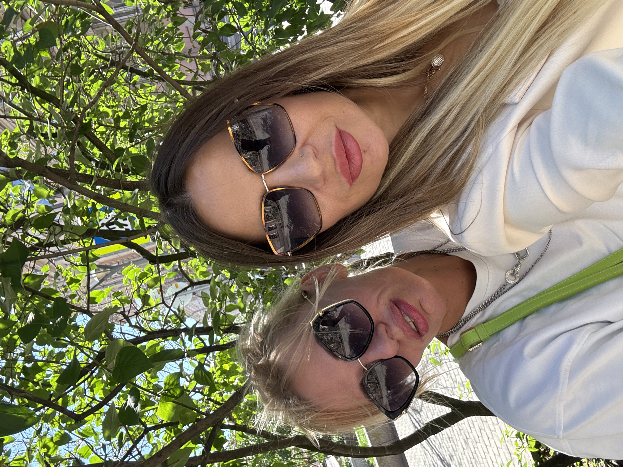 The image is a selfie taken by two women outdoors, possibly in Saint Petersburg, Russia, as suggested by the location context. They are positioned close together, with their faces angled towards the camera. Both women are wearing sunglasses and have makeup on. The woman in the foreground has blonde hair and is wearing a white top with a lime green strap visible. The woman in the background has dark hair and is also wearing a white top. Both are wearing jewelry. They appear to be enjoying themselves, with smiles visible on their lips. The background consists of lush green foliage from trees, with some sunlight filtering through the leaves, indicating it is daytime and likely fair weather. The overall scene suggests a casual outing or a moment of leisure. There is no visible text in the image.