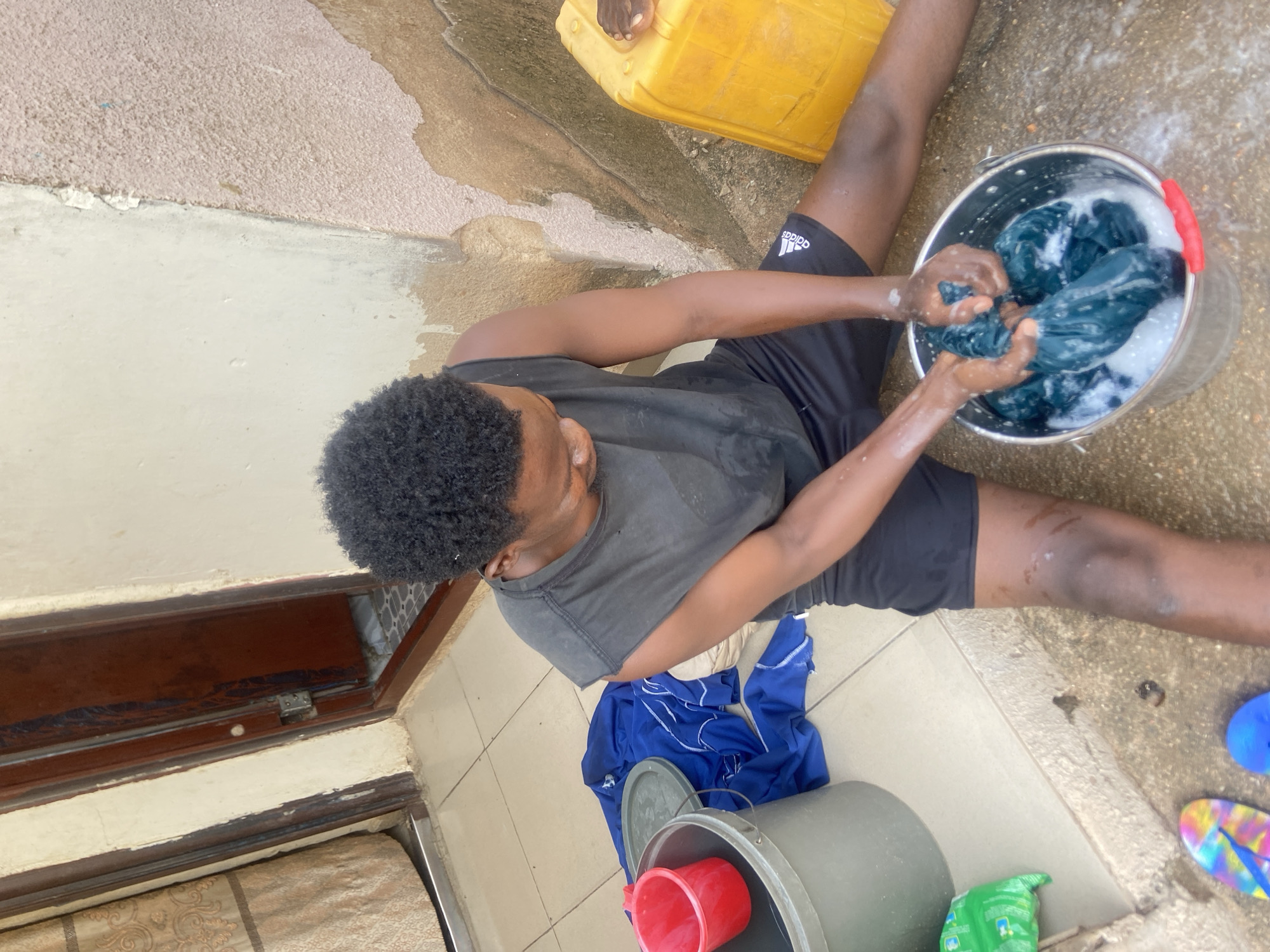 An adult male is observed performing laundry by hand in an outdoor residential area in Jalingo, Nigeria. The individual, dressed in a dark grey sleeveless shirt and black Adidas shorts, is seated on a light-colored tiled patio or porch floor. He is actively squeezing water from dark blue fabric immersed in a silver metal bucket containing soapy water. To his left, another grey bucket with a red plastic cup on top is visible, along with a blue garment laid out on the tiles and a green packaged item, possibly detergent. To his right, a large yellow plastic jerrycan rests against a textured wall showing signs of wear, peeling paint, and discoloration. The floor transitions from light tiles to a rougher, darker concrete surface where the jerrycan is located. In the background to the left, a partially open brown wooden doorframe reveals patterned floor tiles inside, and a beige patterned curtain is visible next to the doorway. The adjacent wall is light-colored and also shows some minor wear. Colorful flip-flops are partially visible on the ground in the lower left foreground.