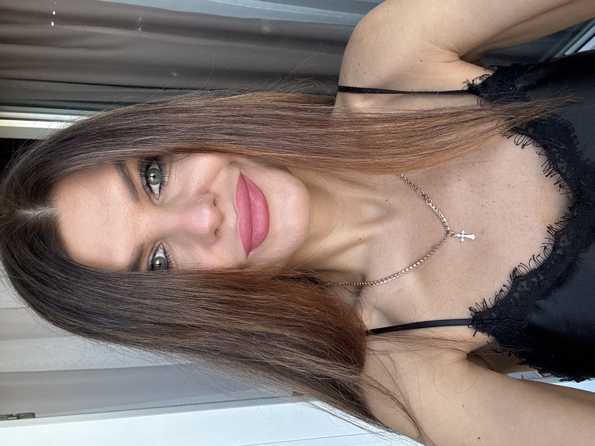 A woman with long, brown hair and green eyes is facing the camera. She has pink lipstick on and is wearing a black lace-trimmed top. A gold necklace with a cross pendant is visible around her neck. The background appears to be indoors, with a window or door frame visible. There is no definitive indication of the location being in Chaikovsky, Russia.