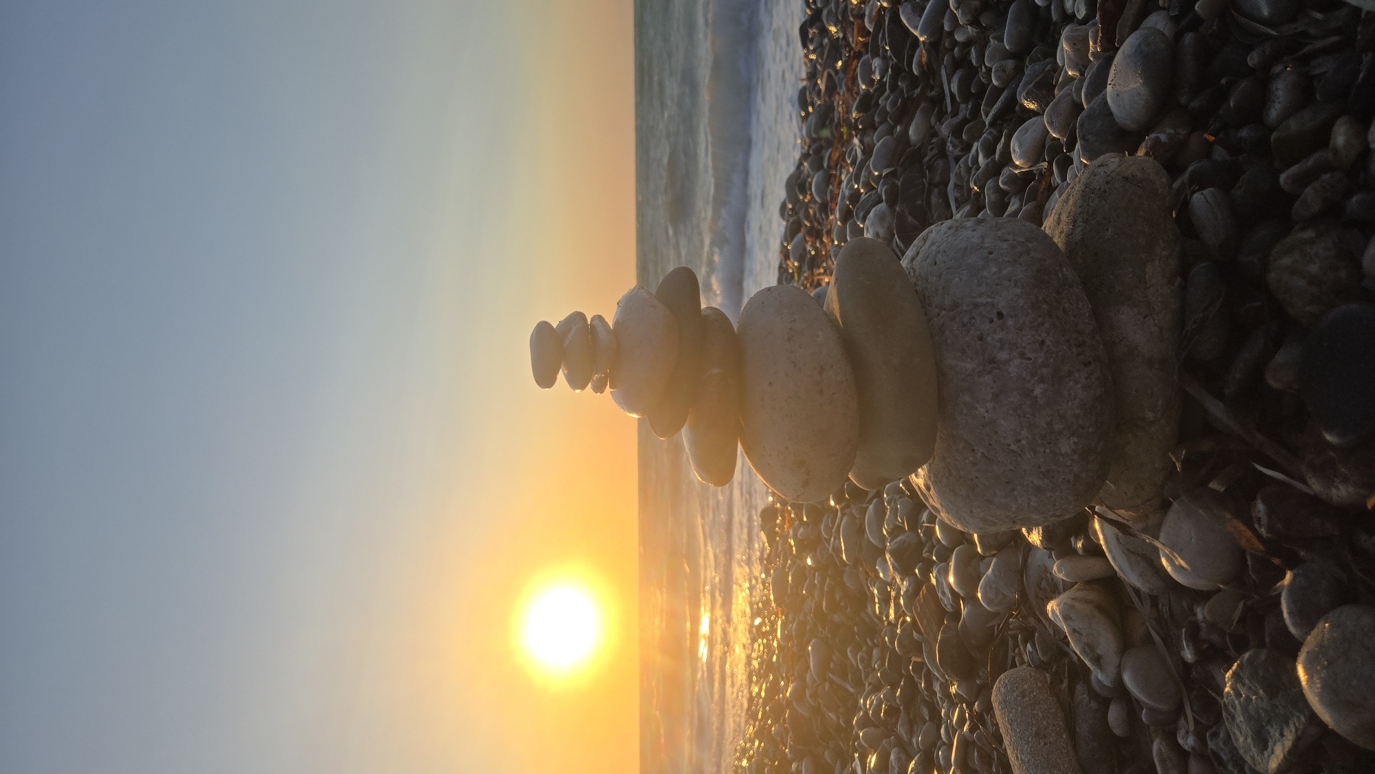 A stacked stone cairn is positioned on a pebble beach at sunset, with the sun, a bright orb of light, low on the horizon and casting a golden glow across the scene. The ocean is visible behind the cairn, with gentle waves lapping at the shore. The sky is a gradient of orange and blue. The beach is covered in smooth, rounded pebbles of various sizes and colors. The cairn consists of several stones stacked one on top of the other, appearing to be balanced precariously. The overall atmosphere is one of tranquility and natural beauty. The location is the beach in Soroni, Greece.