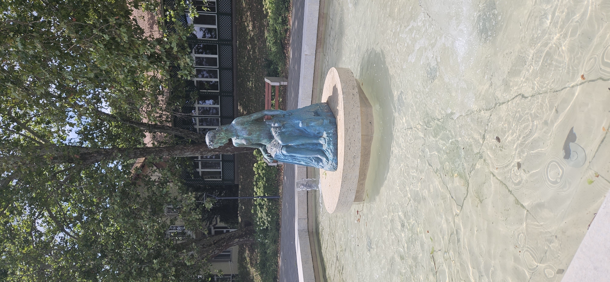 The image shows a bronze statue of a woman sitting in a fountain. The statue is situated on a circular stone base in the center of the fountain, which has clear, shallow water. The woman appears to be partially draped in fabric, and there is a smaller figure, possibly an animal, next to her. Water is gently spraying from the fountain. In the background, there are lush green trees and a building with large windows and a red bench in front of it. A lamppost is also visible. The scene suggests a public park or garden setting. This statue and fountain are located in the town of Hévíz, Hungary, known for its thermal lake.
