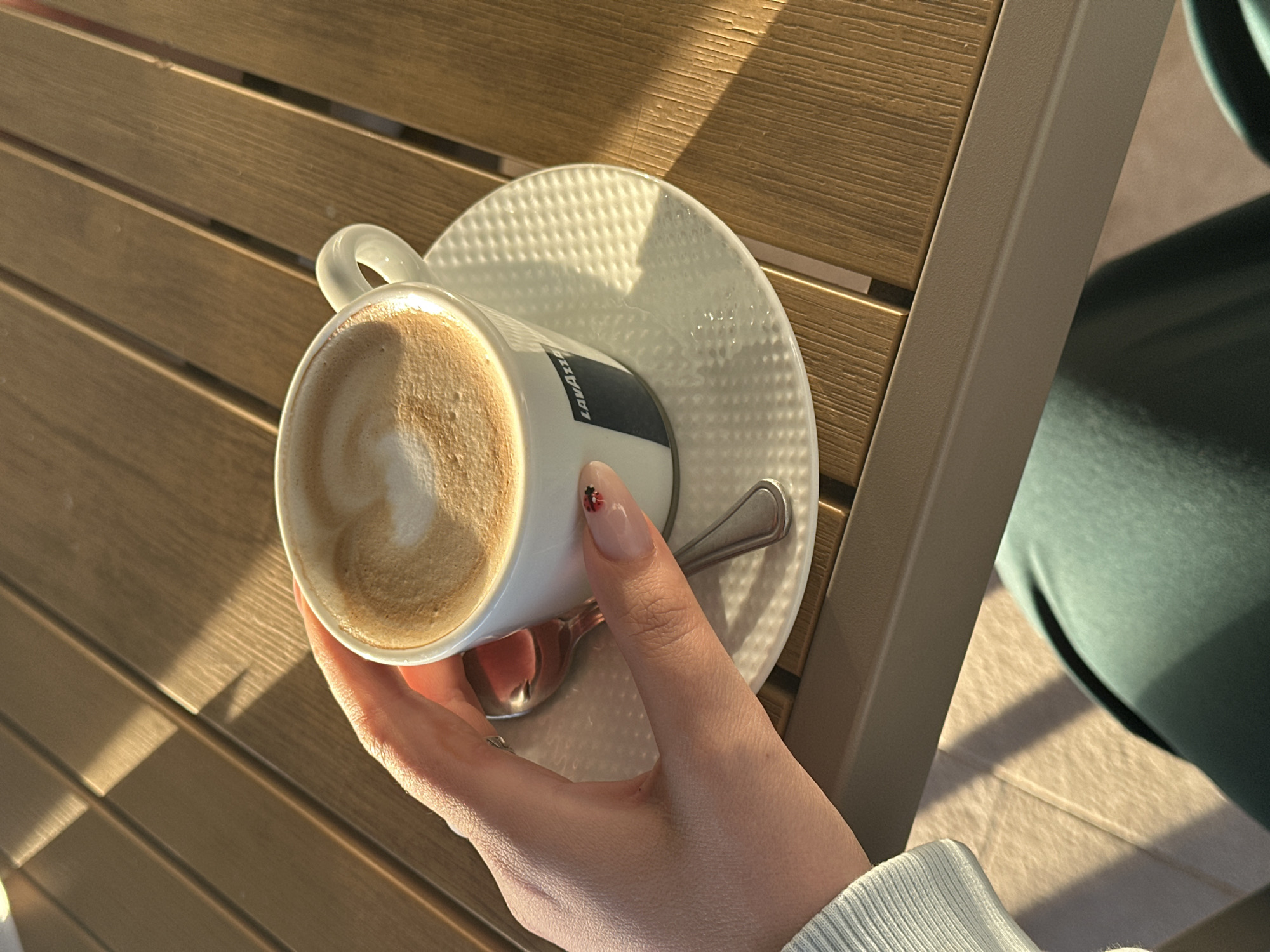 A close-up, high-angle shot captures a person's hand holding a coffee cup on a wooden slatted table, likely outdoors in Bar, Montenegro. The scene is bathed in warm, golden sunlight, indicating either early morning or late afternoon, which casts distinct shadows across the table surface.
The central subject is a white ceramic coffee cup, filled with a light brown, frothy beverage, characteristic of a cappuccino or latte. The brand name "LAVAZZA" is visible in dark lettering on the side of the cup. It sits on a matching white ceramic saucer, distinguished by a unique raised, dotted texture. A silver spoon rests on the saucer.
A fair-skinned hand firmly holds the cup, with the person's wrist partially visible, adorned with a light-colored, ribbed sleeve. Notably, one fingernail has a small, detailed ladybug design in red and black. The table features light brown wooden slats with visible grain. To the right of the frame, a muted green fabric, possibly part of clothing or seating, is partially visible in the background, adding a soft hint of another presence or object. The strong sunlight and shadows contribute to a serene and relaxed atmosphere.