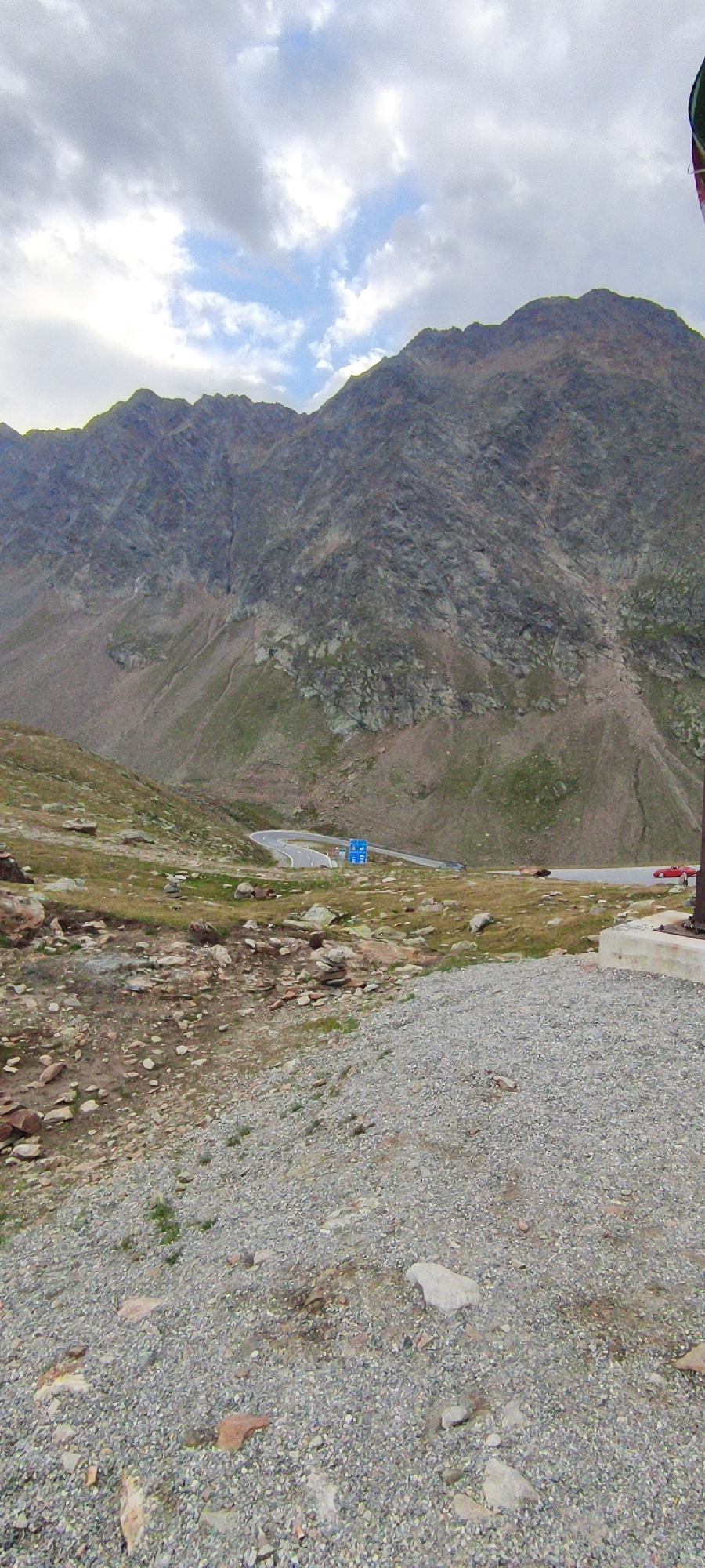 The image depicts a mountain landscape in Sölden, Austria. The foreground is covered with gravel and scattered rocks. A winding asphalt road snakes through the midground, with a few signs visible along its edge. The road is bordered by grassy slopes dotted with rocks. In the background, a large, rugged mountain dominates the scene, its rocky slopes covered in patches of green vegetation. The sky above is partly cloudy, with patches of blue sky visible. A red sports car is parked on the side of the road. The scene appears to be a scenic viewpoint along a mountain pass.