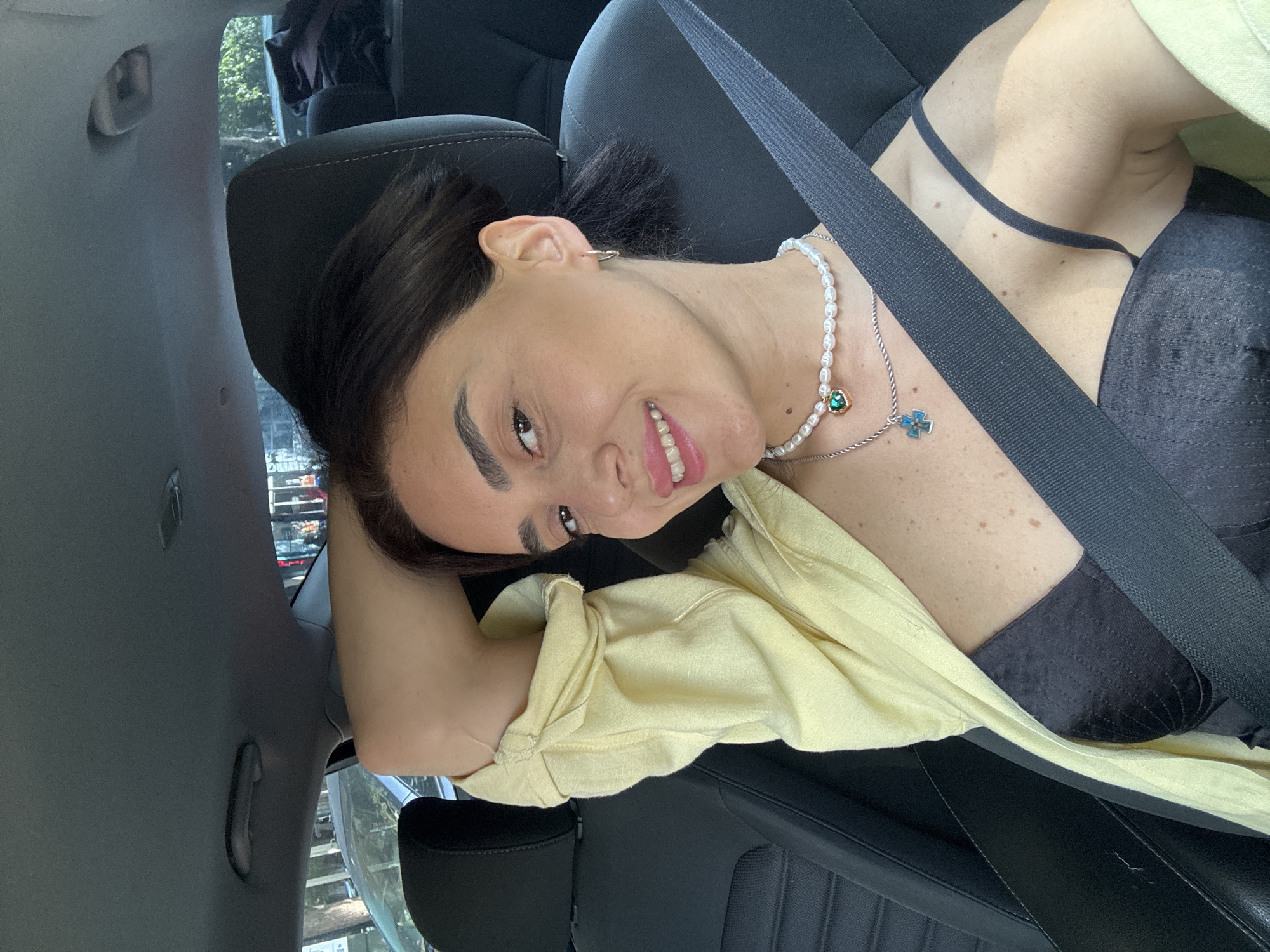 The image shows a woman sitting in the driver's seat of a car. She is smiling and looking at the camera, with her right arm resting on her head. She is wearing a black bustier, a yellow open shirt, and two necklaces. The car appears to be parked on a street in Tbilisi, Georgia, with trees and buildings visible in the background. The lighting suggests it is daytime.