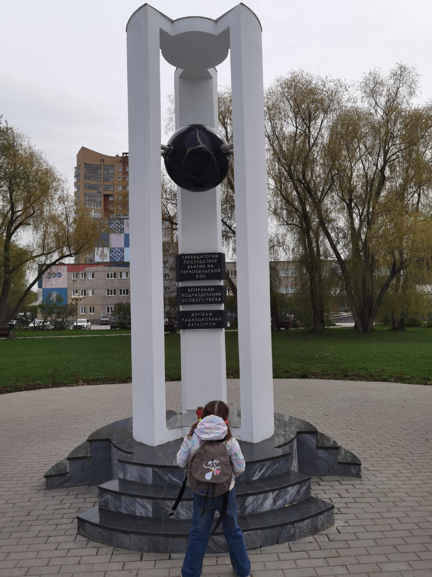 A girl with a backpack stands in front of a monument in Perm, Russia. The monument features three white columns supporting a sphere. Black plaques with white Cyrillic text are affixed to the monument. The monument is elevated on a multi-tiered stone platform. The area around the monument is paved with bricks and surrounded by a lawn, trees, and buildings. The sky is overcast.