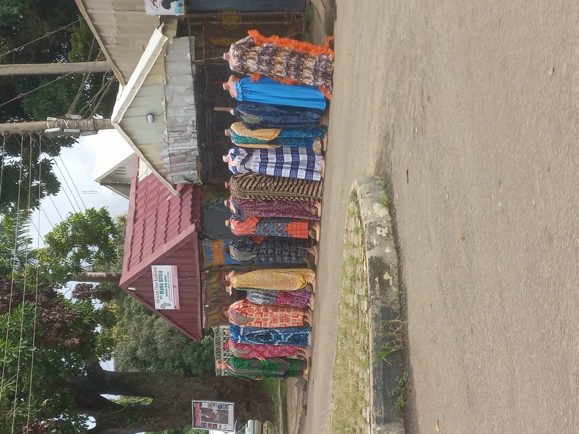 A row of mannequins, adorned in colorful, patterned African attire, are displayed outside a shop in Jos, Nigeria. The shop, characterized by a red-tiled roof and corrugated metal siding, appears to be selling fashion and arts and crafts. A sign on the shop reads "African Fine Fashion Mama Africa - Women & Kids Wears, Arts & Crafts Work." The mannequins are arranged on the pavement, with a tree on the left side of the image and overhead utility wires and poles visible. The scene is set on an asphalt road with a concrete curb and some overgrown grass. The overall ambiance suggests a commercial street scene in a Nigerian city.