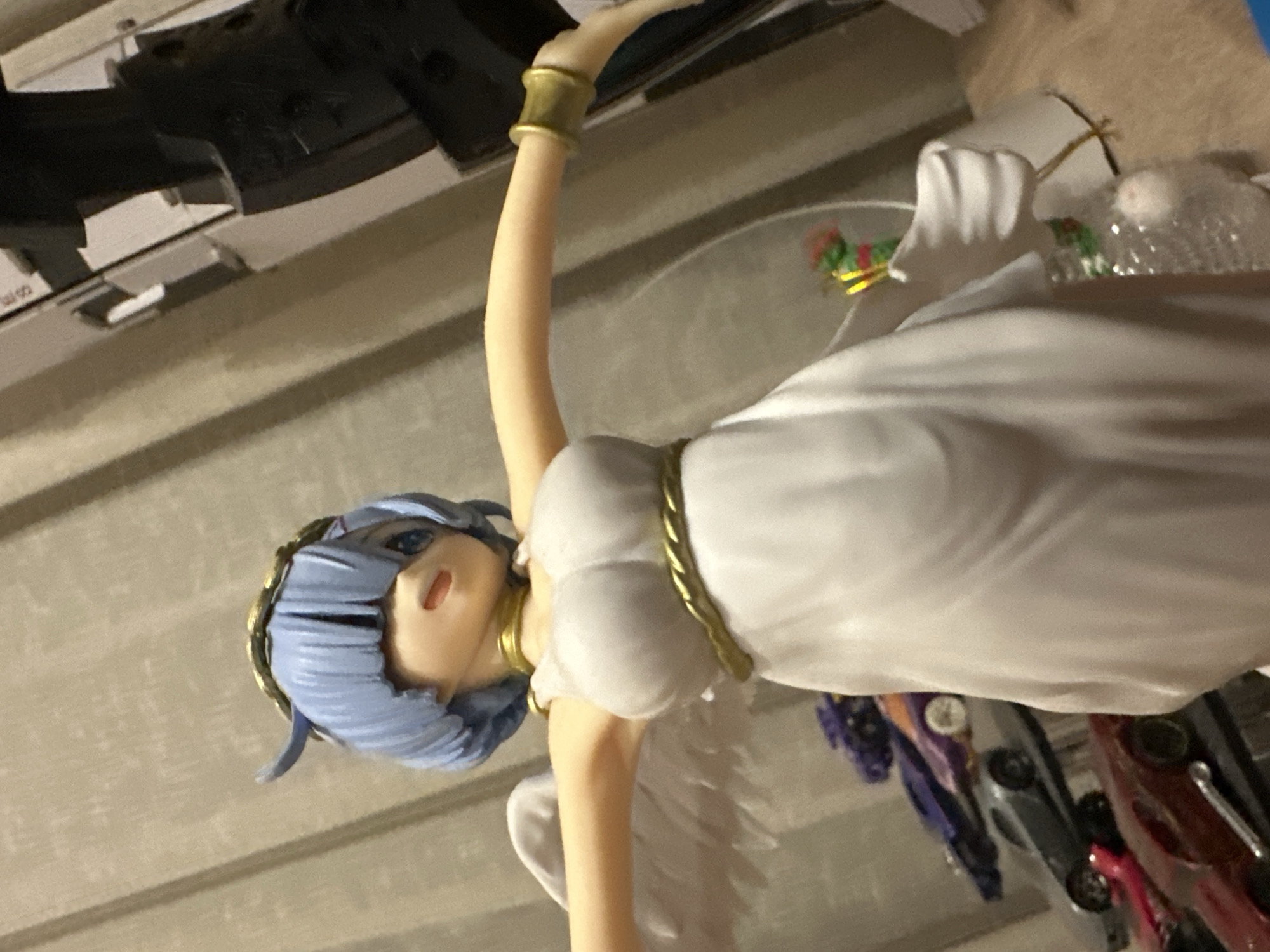 The image depicts a close-up, waist-up shot of a female figurine with short, light blue hair, large blue eyes, and light skin. The figurine is wearing a white, flowing dress with a gold ribbon around its waist and a golden halo or crown on its head. It has white feathered wings extending from its back. The figurine's arms are outstretched to the sides. In the background, on the left, several toy cars are visible, stacked on shelves. The figurine is positioned in front of a beige, vertical-striped curtain. The lighting is dim and appears to be artificial. The overall setting is indoors, with the figurine being the primary subject.
