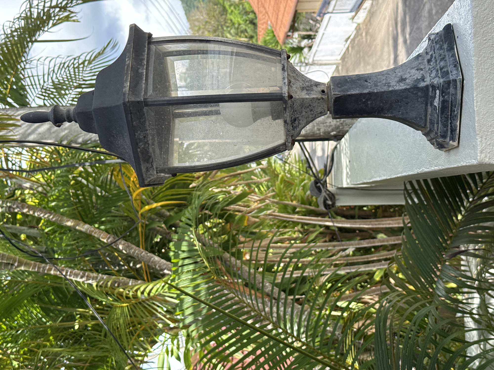 An eye-level, outdoor shot depicts an antique, black, glass-encased electric lamp affixed to a white, exterior wall in Rawai, Thailand. The lamp features a decorative finial at the top and is visibly weathered, with spots and dust on the glass. Lush tropical foliage, including palm fronds, surrounds the lamp, obscuring the background partially. Electrical wires are visible amidst the foliage. The sky is partly cloudy. A portion of a red tiled roof is seen behind the white wall.