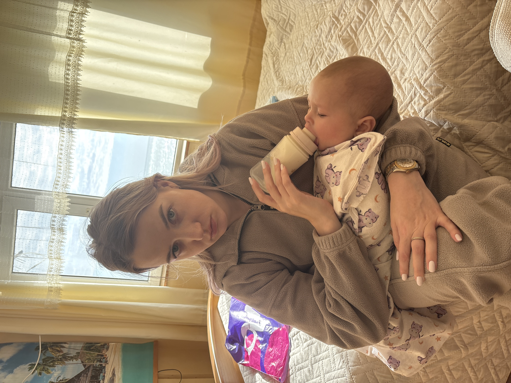 A woman is sitting on a bed, feeding a baby from a bottle. The woman is wearing a brown fleece jacket and has long blonde hair. She is holding the baby in her arms, supporting its head with her left hand while feeding it with her right. The baby is wearing a white pajama set with purple animal prints.
The bed has a white quilted bedspread. In the background, there is a window with sheer white curtains and a view of the sky. There is a painting on the wall with palm trees. The word "NEVERLATE" is visible on the sleeve of the woman's jacket. A purple and pink bag is on the bed as well. The location cannot be determined as Sochi, Russia.