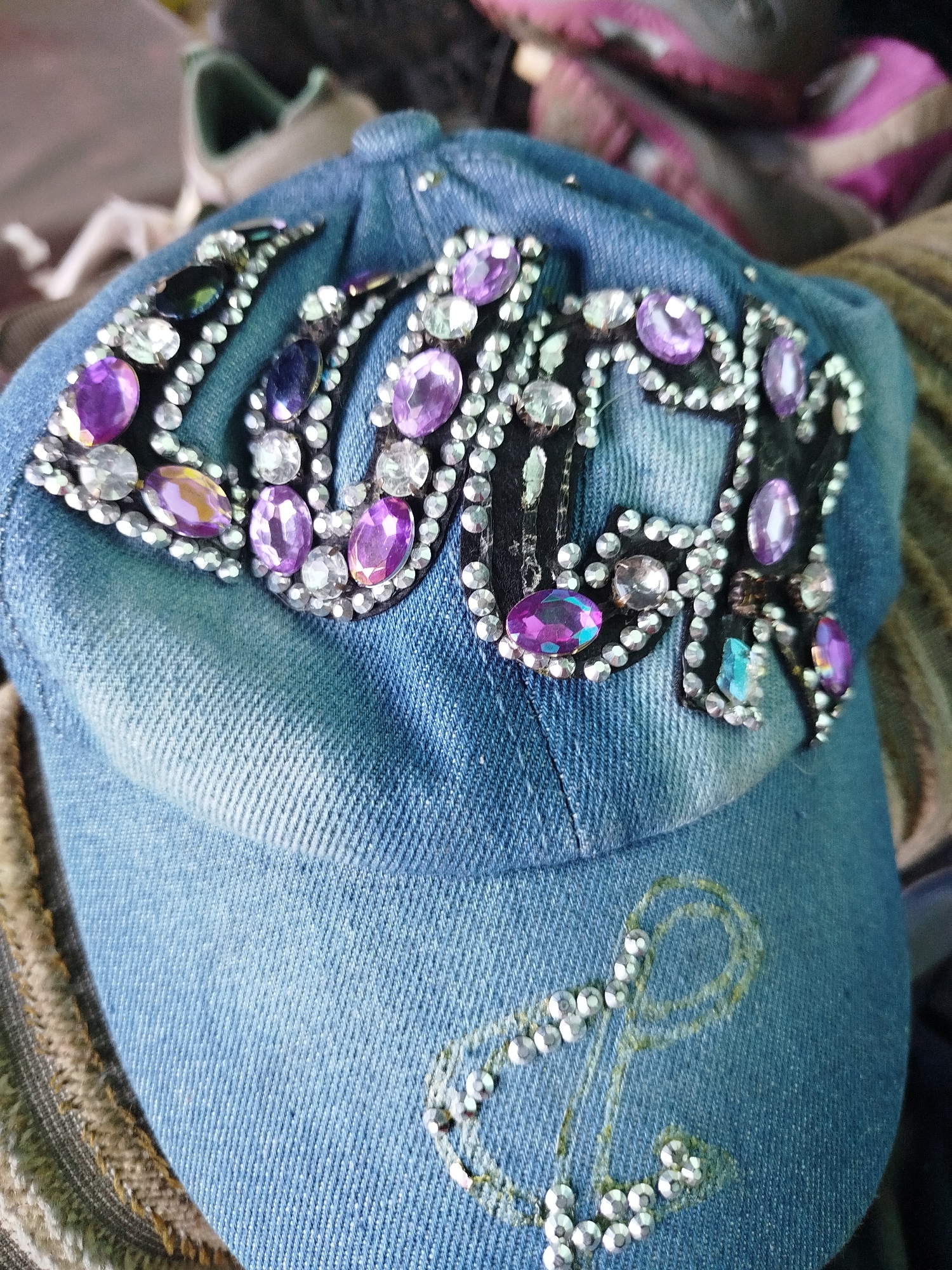 A close-up, top-down view of a denim baseball cap adorned with rhinestones and purple embellishments that form the word "LOVE." The cap is a light blue denim material and is decorated with an anchor shape made of rhinestones on the side. The background is blurred, showing what appear to be shoes and fabric. The scene is indoors and does not provide any information about the location in Крывасельскі сельскі Савет, Belarus. There are no people present in the image.