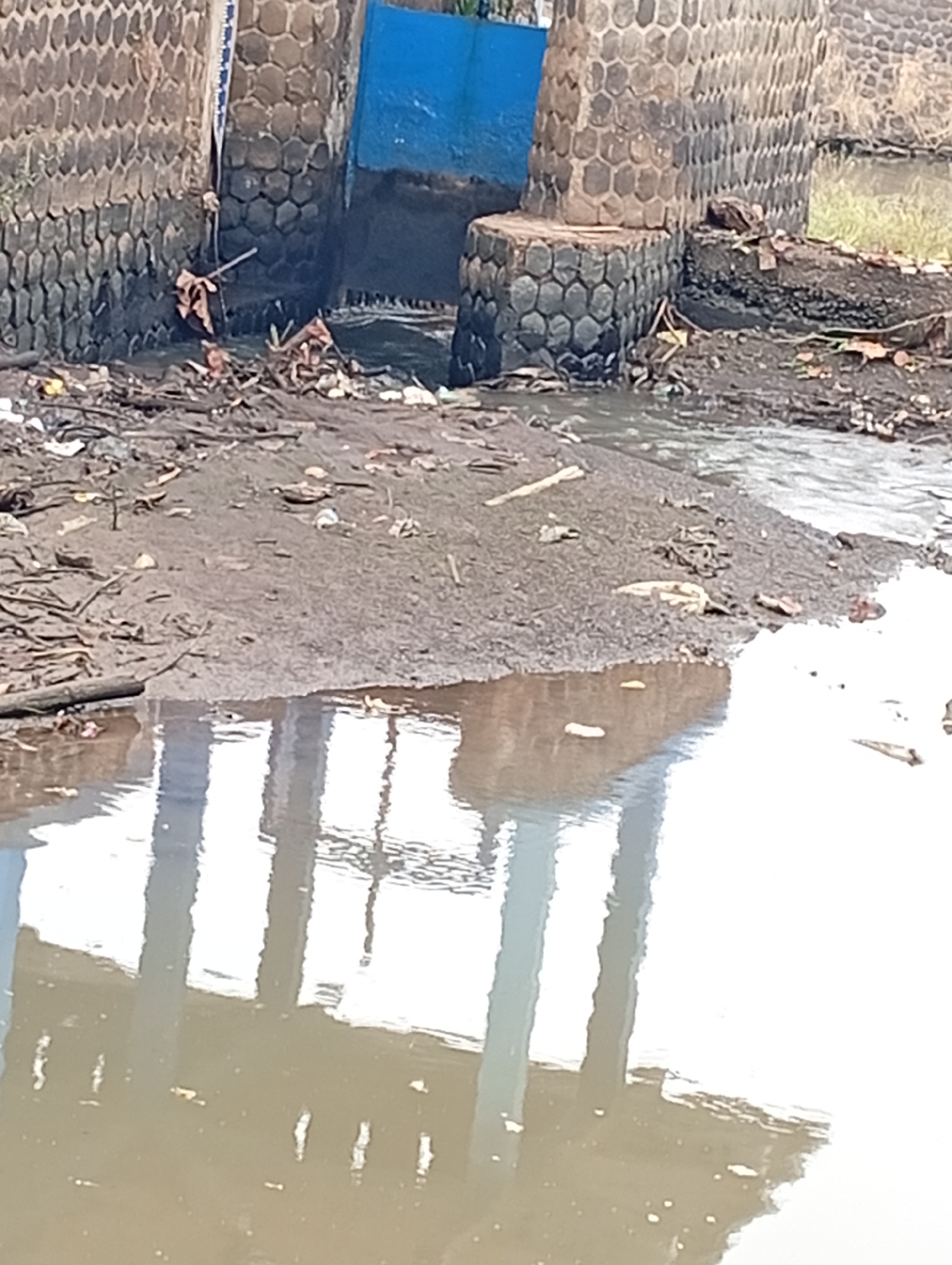 The image depicts a scene of environmental neglect. A concrete structure with a blue opening is visible, suggesting a sluice gate or water passage. The surrounding area is heavily littered with debris, including plastic waste, organic matter, and branches. The ground appears muddy and uneven. The water body in the foreground is murky, with reflections of structures that are not fully visible. The overall impression is one of disrepair and pollution in an outdoor urban or semi-urban environment.