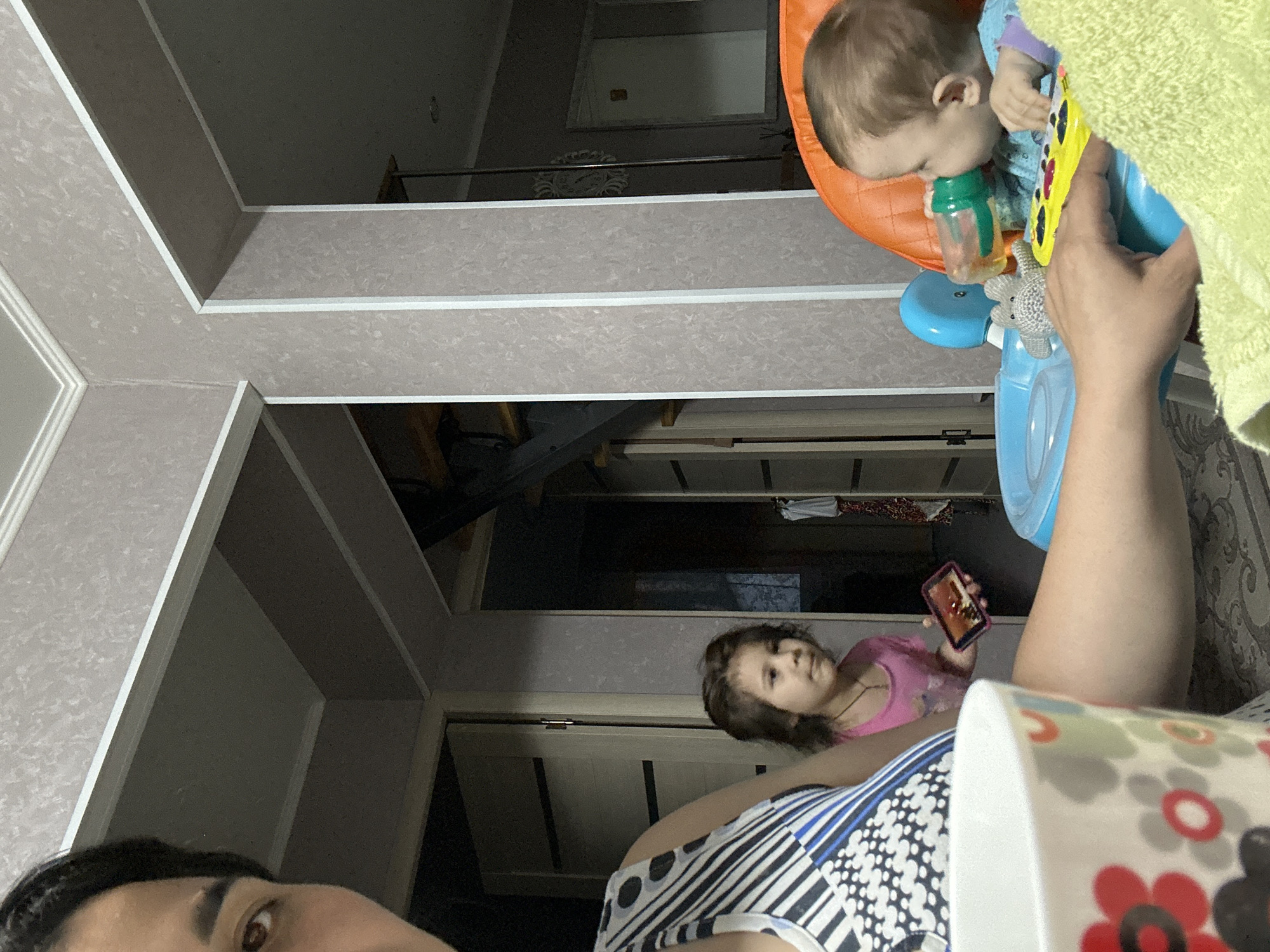An intimate, low-angle indoor scene features three individuals: an adult and two children. In the bottom left, the right eye and part of the forehead of an adult are visible, suggesting a self-portrait or close-up shot from a prone position.
In the upper right, a baby with light brown hair is seated in an orange and blue bouncer or activity seat. The baby is drinking from a green bottle and holds a yellow toy, with a small grey stuffed animal attached to the seat. A green towel is partially visible near the baby.
Towards the middle right, an older child with brown hair is lying down, wearing a pink top, and holding a pink smartphone. A white mug with red, black, and white floral patterns is partially visible in the bottom right foreground.
The background reveals a residential interior with patterned light-colored wallpaper and a distinctive ceiling structure featuring recessed panels or beams with white trim. Several doorways, a mirror or framed artwork on a distant wall, and a decorative white wall hanging or clock behind the baby seat are also visible.
The lighting is ambient, suggesting an indoor setting. The atmosphere is relaxed and informal, capturing a candid family moment. No visible text is discernible in the image.