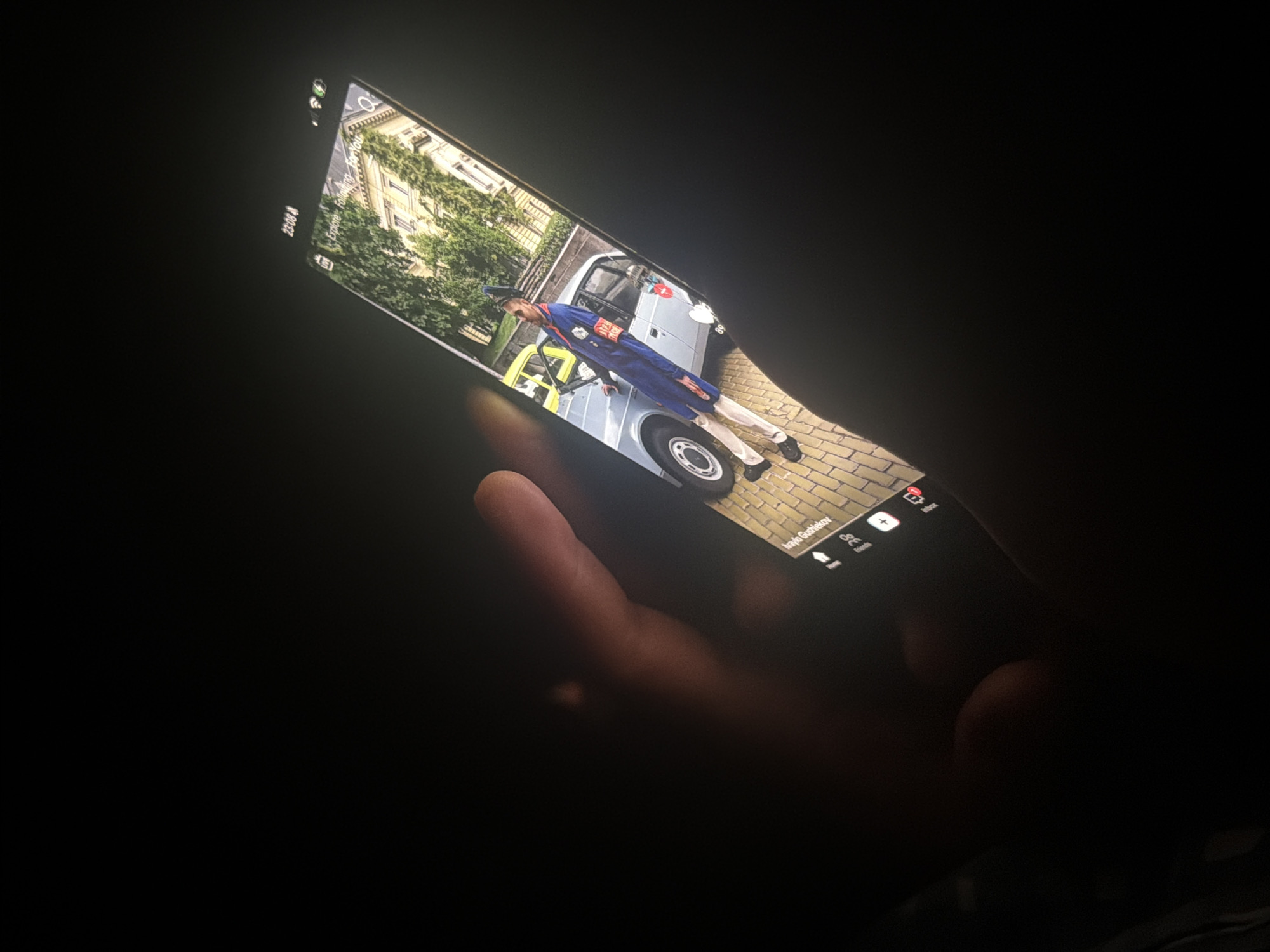 A hand, with fingers visible, holds a smartphone in a dark environment. The phone's screen is brightly illuminated, serving as the primary light source in the image. The time displayed on the phone is 23:08.
The phone screen displays an image featuring a person in what appears to be a uniform. The individual wears a dark blue jacket with a red armband, light-colored trousers, black shoes, and a peaked hat. They are standing next to a light grey car or van, which has a distinct yellow or light-colored railing near its front. Behind the vehicle and the person, there's a multi-story building with architectural details and green trees. The ground beneath the person and vehicle is paved with cobblestones.
Additional text and interface elements are visible on the phone screen: "Explore", "Following", "For You" at the top, and "Home", "Friends", and the name "Ivaylo Gushlekov" at the bottom, along with an "Add" button and a partially visible word that seems to end in "book". A battery and Wi-Fi icon are also discernible in the top left corner. The image on the phone appears to be from a social media or content-sharing platform.