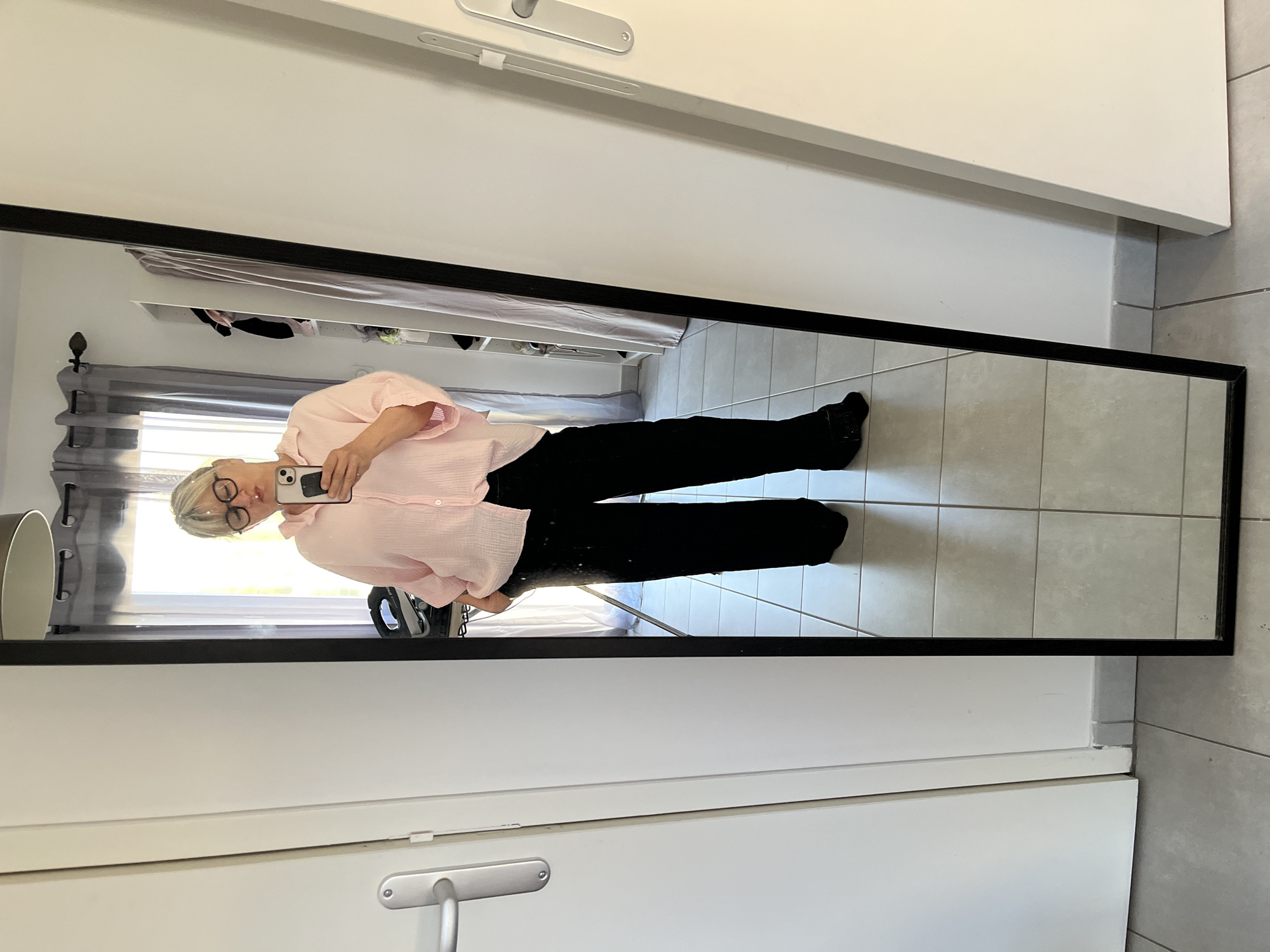A person stands in front of a full-length mirror, holding a smartphone and taking a picture of their reflection. The person is wearing a light pink collared shirt, black pants, and black socks. The mirror is framed in black and is hanging on a white wall. The background includes a partially visible room with a curtained window, shelving, and a gray tiled floor. The location is identified as Chambœuf, France, based on available information.