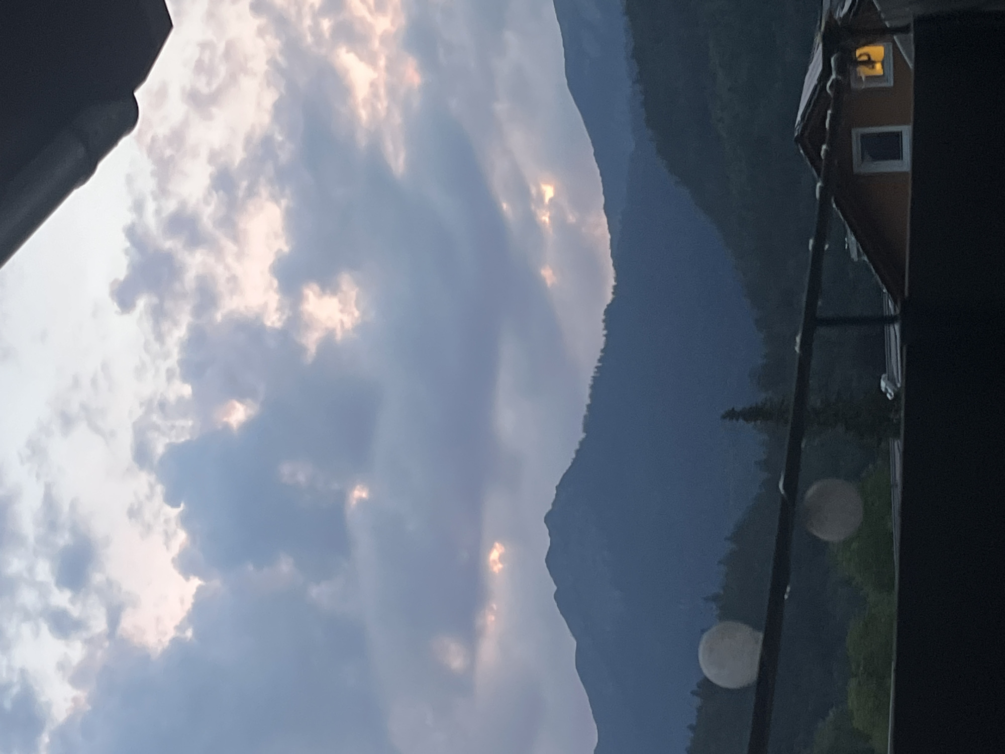 The image depicts a mountainous landscape, likely taken from an elevated viewpoint, possibly a balcony or rooftop, overlooking the town of Garmisch-Partenkirchen, Germany. The foreground features a dark railing with two illuminated spherical objects, possibly lights or decorative orbs, suspended by thin wires. Beyond the railing, the scene transitions to a densely wooded hillside. In the middle ground, the silhouette of a building with a sloped, dark roof is visible, featuring a lit window emitting a warm yellow glow. Further into the distance, majestic mountains rise, their peaks partially obscured by a dramatic, cloudy sky. The clouds are tinged with soft pink and orange hues, suggesting either sunrise or sunset, with shafts of light breaking through in places. The overall impression is one of a serene, twilight scene in a mountainous region.