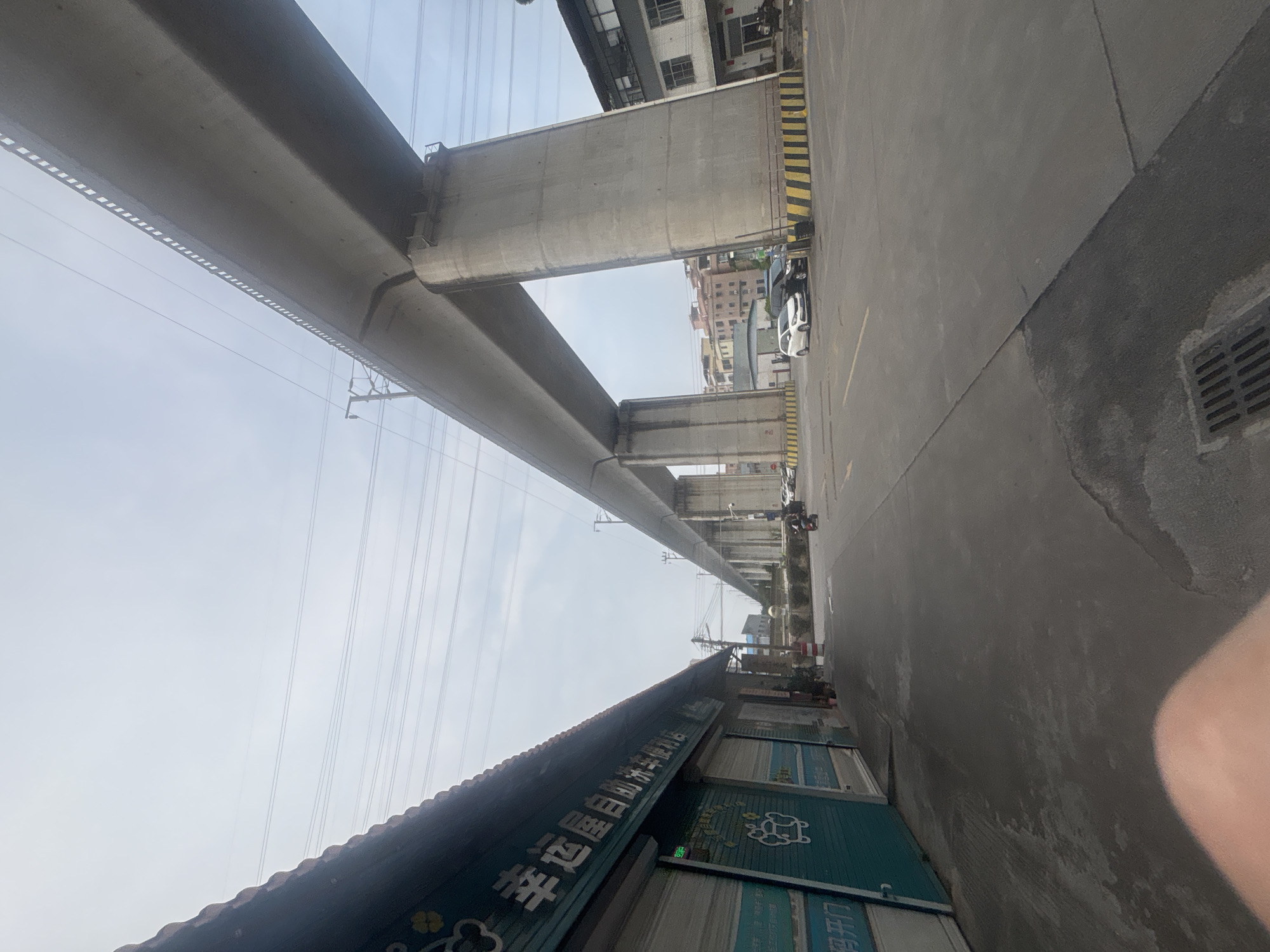 A wide-angle, eye-level shot captures an outdoor scene in Nanhai District, China, dominated by a massive elevated highway structure. The highway, constructed of concrete, spans across the frame supported by thick, imposing pillars. Below the highway, on the left side of the image, is a row of storefronts with teal-colored rolling security gates, some adorned with text and cartoonish graphics. One gate displays Chinese characters and a whimsical image of a paw print. To the right of the storefronts, a paved road leads towards the highway pillars. A few cars are parked along the side of the road, and a motorcycle with a rider is visible in the middle distance, moving away from the camera. The environment appears to be urban or semi-urban, with buildings and infrastructure typical of a developed area. The sky above is overcast and pale.