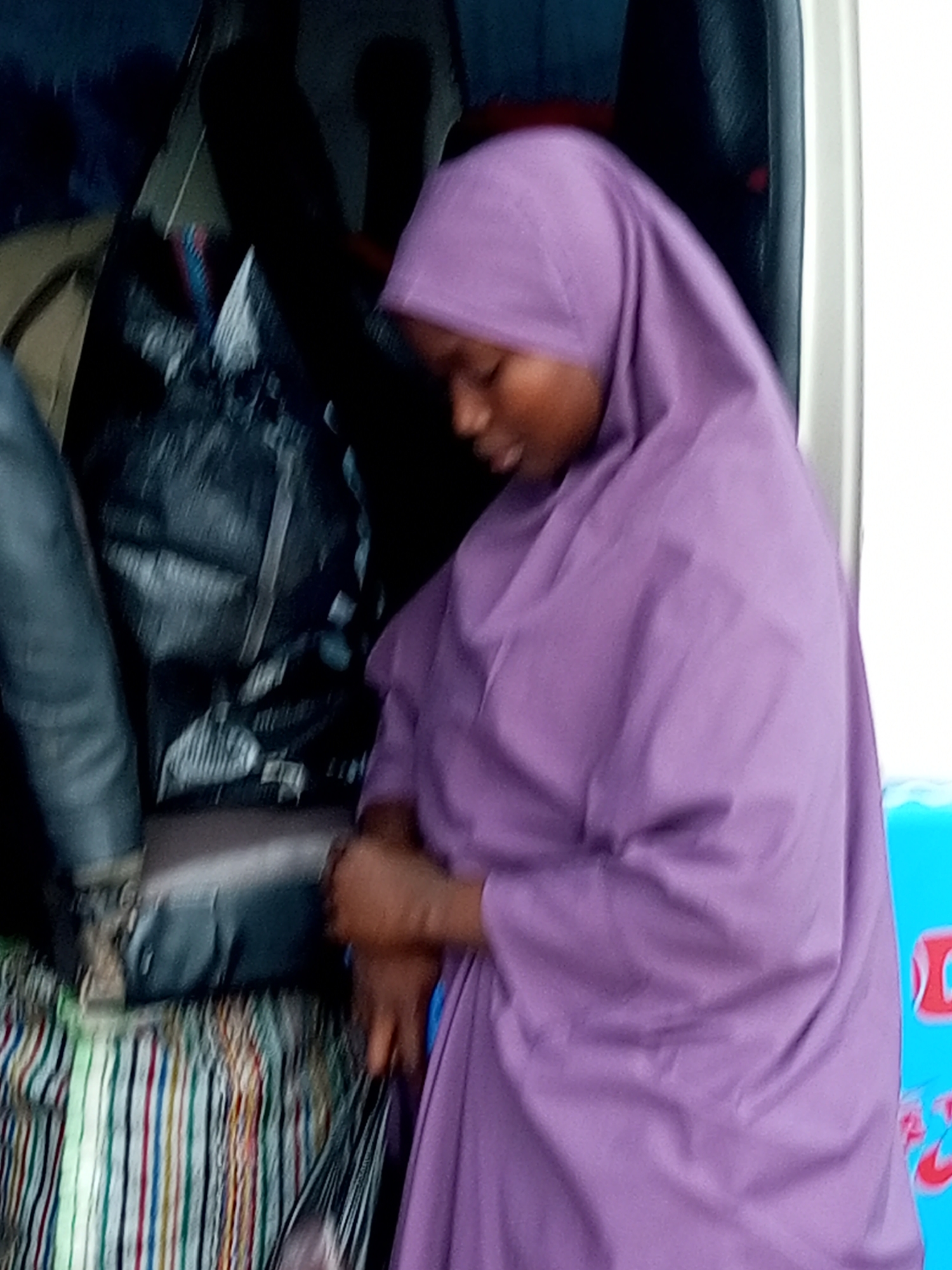 In Gombe, Nigeria, a person is captured in an indoor setting, likely a vehicle, wearing a purple head covering and a matching outfit. They are seated and appear to be engaged in an activity with their hands, possibly packing or organizing belongings. The background is cluttered with bags, including a large black duffel bag, and a striped fabric is visible beneath. The lighting suggests an overcast day or an indoor environment. The scene conveys a sense of transit or preparation for a journey.