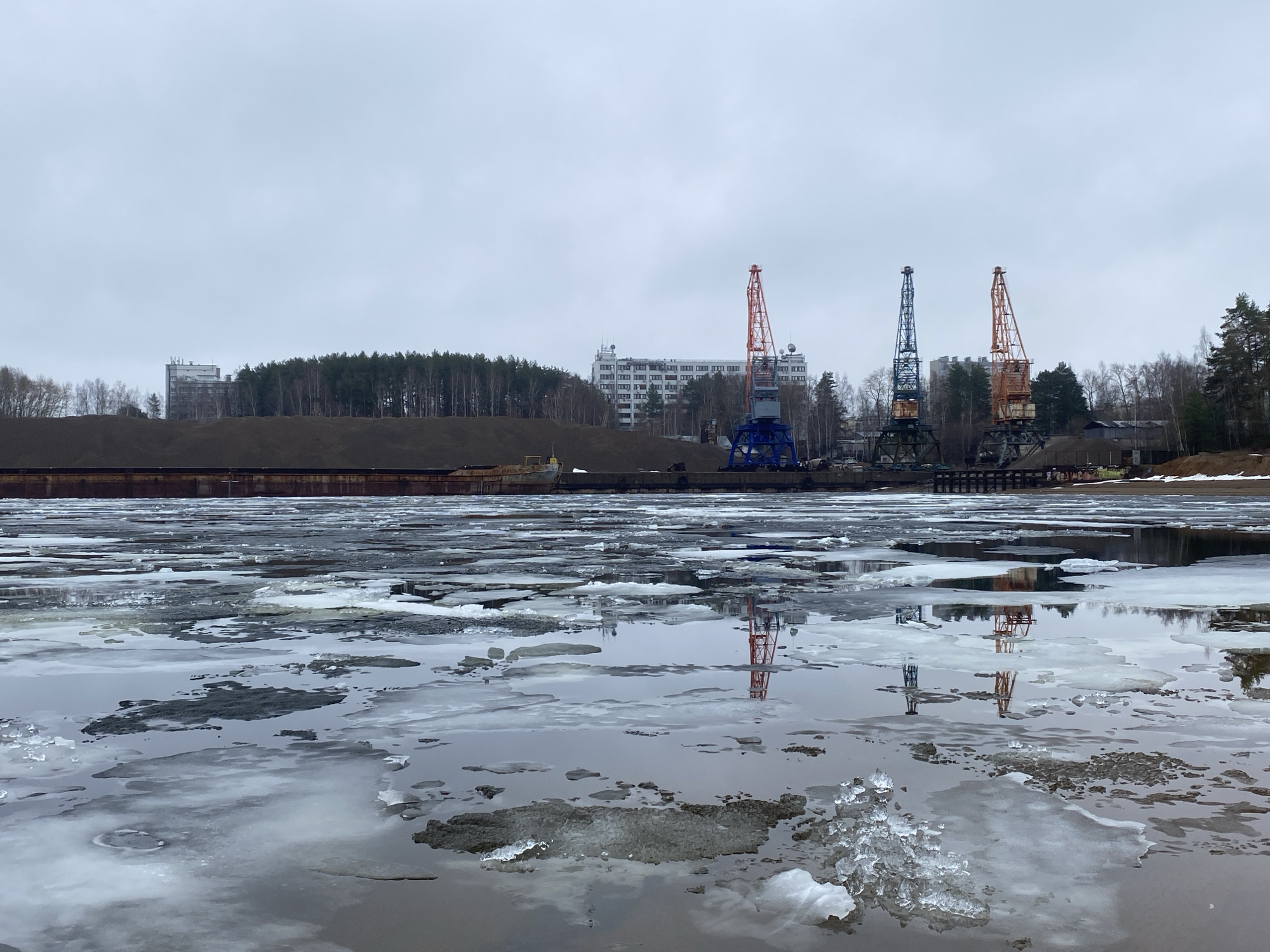 The image displays an industrial waterscape in Chaikovsky, Russia, under a consistently overcast, light grey sky. In the foreground, a body of water, likely a river or canal, is largely covered by numerous broken ice floes and patches of slush, suggesting a cold environment or the onset of a thaw. The still water between the ice shards creates clear reflections of the cranes and the cloudy sky above.
The midground features a rugged shoreline, where a large, dark brown mound of bulk material, possibly sand or gravel, is piled. Three prominent gantry cranes stand along the water's edge, indicating an active port or cargo handling facility. The cranes are distinct in color: one is blue with a black base, while the other two are brightly colored in orange and red.
Behind the cranes and the material pile, a dense band of trees, comprising both bare deciduous and evergreen conifers, stretches across the horizon. Several buildings are visible through and above the trees, including a large, multi-story white building with antenna-like structures on its roof, positioned towards the left-center.
The scene appears devoid of active human presence or ongoing work, lending it a quiet, somewhat stark industrial atmosphere. The diffused light suggests daytime, though the heavy cloud cover prevents direct sunlight. No discernible text is visible within the image.