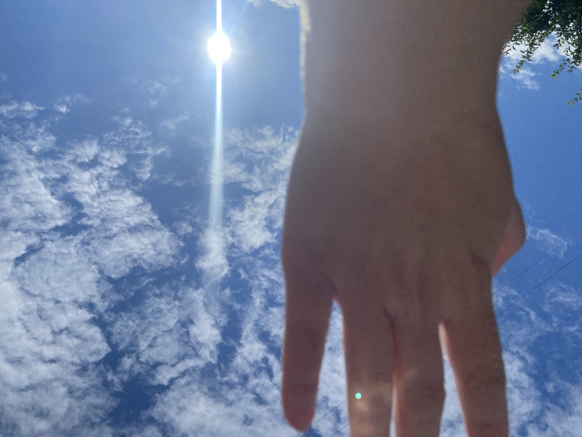 A low-angle shot captures a person's hand reaching upwards against a bright blue sky filled with scattered white clouds. The sun is a brilliant orb with rays of light emanating from it, creating a lens flare effect. The hand, partially out of focus, obscures some of the sky. In the upper right corner, a portion of green foliage is visible. The scene appears to be outdoors, under a sunny sky. The location is Askarovo, Russia.