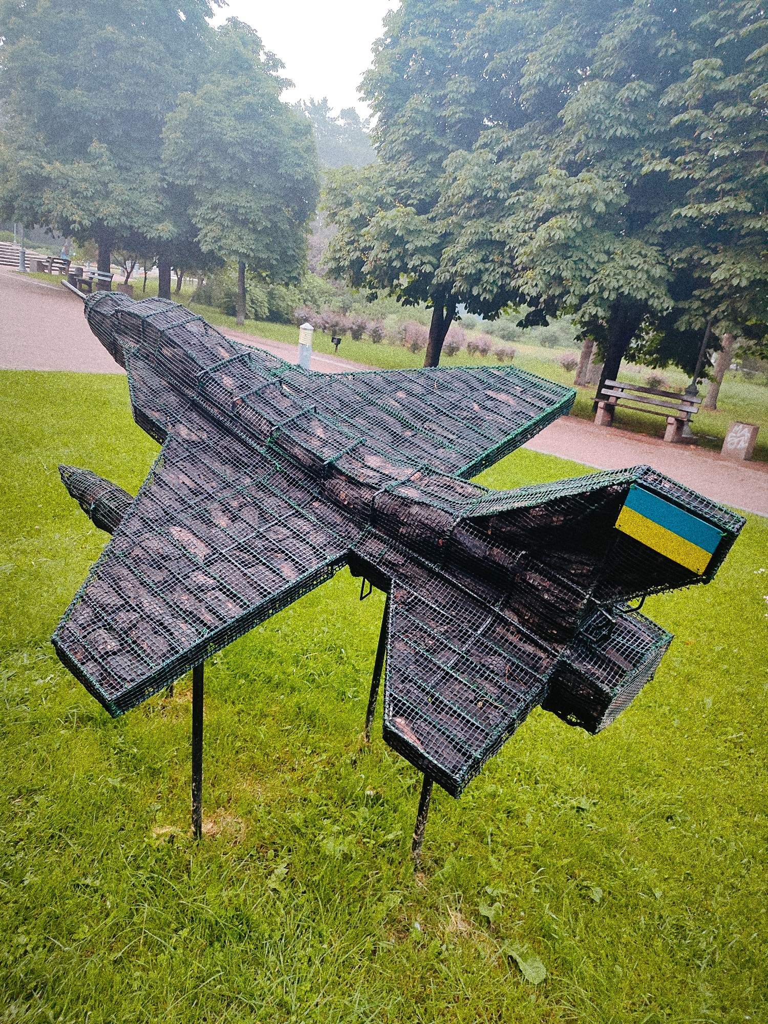 The image displays an outdoor scene in Kyiv, Ukraine, centered on a large, abstract sculpture of a fighter jet. The sculpture is primarily constructed from dark, irregularly shaped organic material, such as wood fragments or branches, encased within a green, rectangular wire mesh that forms the aircraft's exterior. The jet's design features twin tailfins and an overall silhouette resembling a Soviet-era fighter. A rectangular panel, horizontally divided into a blue upper section and a yellow lower section, representing the flag of Ukraine, is affixed to the outer side of the sculpture's right tailfin.
The sculpture is supported by at least two dark, slender posts embedded in the ground, elevating it above a vibrant green grassy lawn. The grass immediately surrounding the support posts shows some lighter, drier patches. In the background, a light-colored paved pathway curves through the landscape, flanked by multiple mature deciduous trees with dense green foliage. Several stone or concrete park benches are visible along the pathways, suggesting a public park environment. The overall lighting is soft and diffused, indicating an overcast sky, with a slight atmospheric haze present among the distant trees. No individuals are visible in the scene.