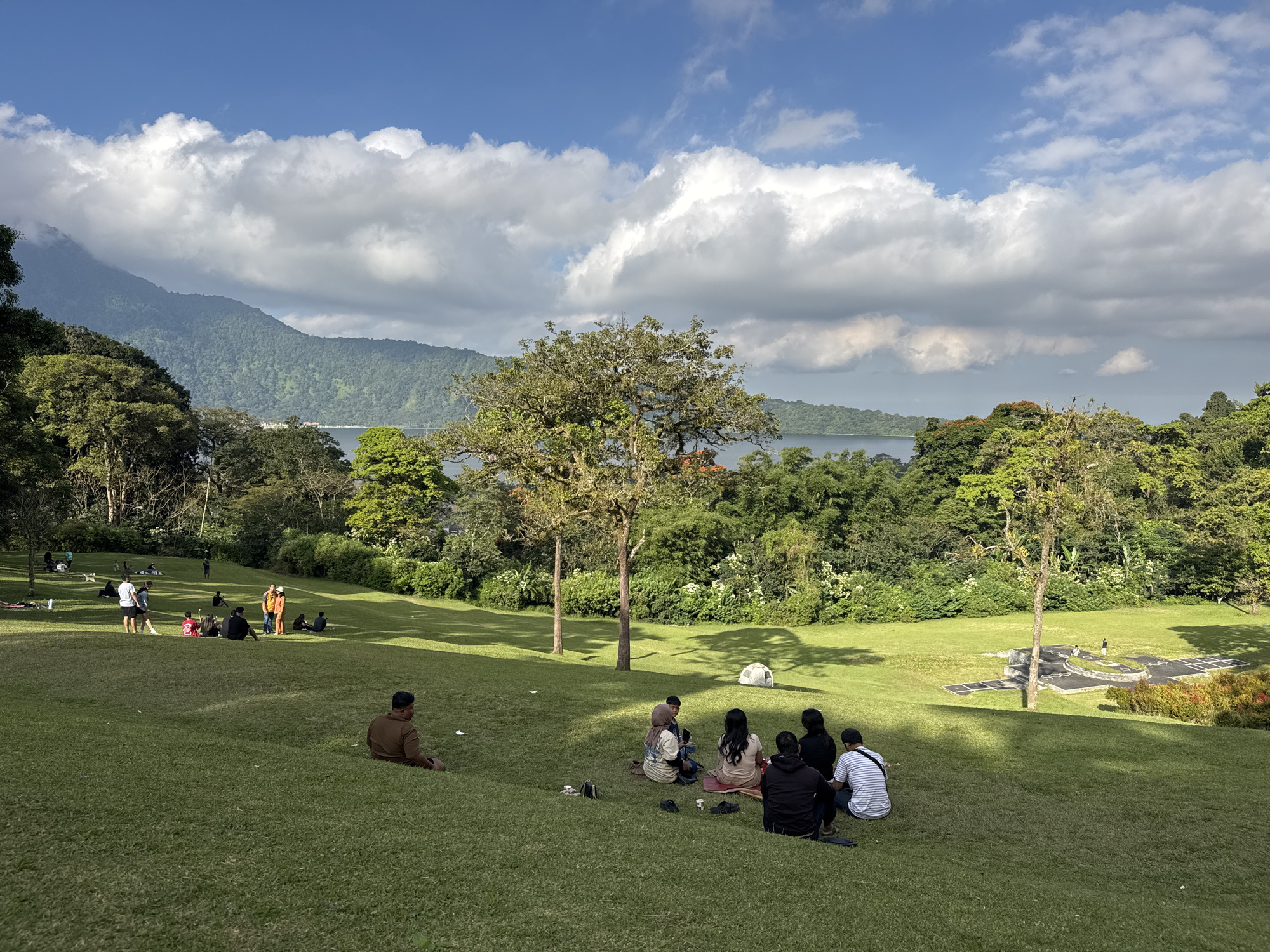 This image depicts a serene outdoor scene in Baturiti, Indonesia, likely during the daytime. A group of people are enjoying a leisurely day on a lush, green grassy hill. Several individuals are seated on the grass, some in small groups, possibly having a picnic or simply relaxing and conversing. In the background, a body of water is visible, bordered by dense, verdant trees and rolling hills, with a large mountain range in the distance under a bright blue sky dotted with fluffy white clouds.
The atmosphere appears peaceful and relaxed, with people casually dressed and engaged in quiet activities. Some are sitting individually, taking in the scenery, while others are gathered together, suggesting a social outing or family gathering. A small white tent is visible in the middle distance, adding to the sense of a recreational outing. The sunlight casts gentle shadows on the grass, indicating it is likely mid-morning or afternoon. The vibrant green of the landscape, the clear blue sky, and the presence of mountains and water suggest a scenic natural location, possibly a park or a viewpoint overlooking a lake or reservoir. There is no visible text in the image.