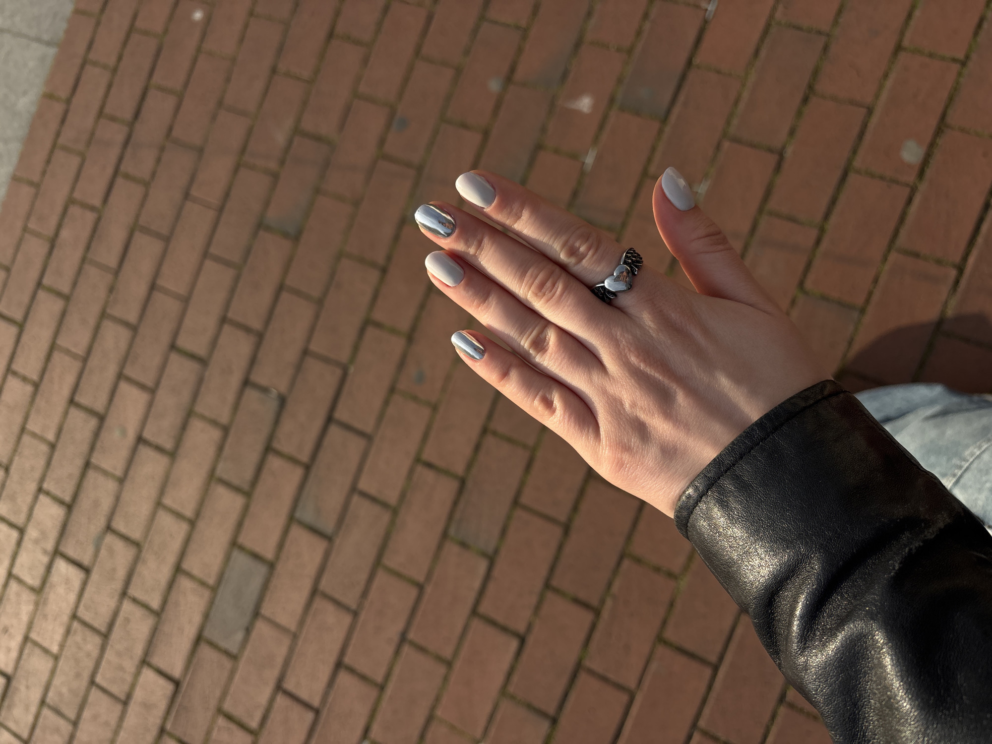 The image provides a close-up, high-angle perspective of a person's right hand, prominently displaying their manicure and a ring against an outdoor setting. The hand is illuminated by bright, directional sunlight, suggesting either early morning or late afternoon.
The subject's nails are neatly filed and polished. Most nails feature a solid, light gray or greige color, while the index finger and ring finger sport a striking metallic silver or chrome finish. A notable silver-toned ring is worn on the middle finger, designed with intricate details that appear to be two interconnected heart shapes or possibly wings.
The person is dressed in a black leather jacket, with the sleeve visible in the lower right quadrant of the frame. A small portion of light blue denim fabric is also discernable in the top right corner, indicating the wearer might be in jeans or another denim garment.
The background consists of reddish-brown brick paving stones, laid in a diagonal pattern. Faint green patches, likely moss or grass, are visible in the crevices between some of the bricks. The strong sunlight casts soft shadows, highlighting the textures of both the hand and the paved surface.
This scene is set in Varna, Bulgaria, capturing a casual moment of personal style in an urban outdoor environment. There is no visible text in the image.