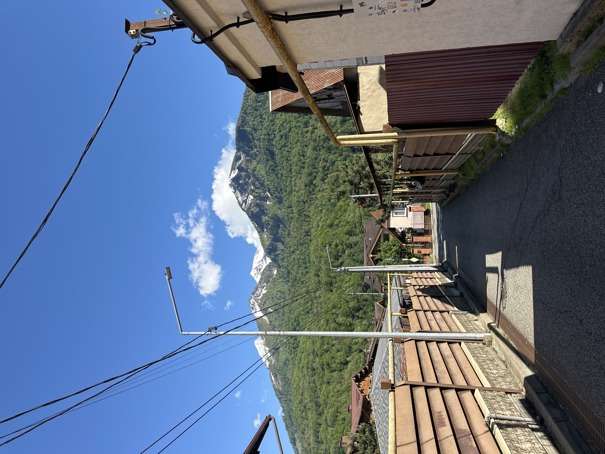 The image presents a scenic view of a narrow street or alleyway in a mountainous settlement, likely Krasnaya Polyana, Russia, given the provided context. The perspective looks down the paved street, which is bordered by buildings on the right and a majestic mountain range in the background.
The buildings on the right are somewhat rustic, featuring different roof types including corrugated metal and shingles. A rusted yellow pipe runs horizontally along the exterior of one structure. Utility poles and a modern street lamp stand along the street, with multiple power lines crisscrossing the bright blue sky overhead. Along the edge of the street, patches of green grass and weeds are visible.
Dominating the background are towering mountains, densely covered in lush green forest, suggesting a verdant, natural environment. Patches of snow are visible on the higher peaks of the mountains, indicating a high altitude or a cool climate. The sky is a vibrant blue with a few fluffy white clouds scattered across it.
The scene is bathed in bright daylight, with strong shadows cast on the street and buildings, indicating a clear, sunny day, likely in the morning or early afternoon. No people are visible, contributing to a tranquil and serene atmosphere. On the underside of an eave of a building in the immediate foreground, a collection of small stickers or graffiti is discernible, though specific text is not clear.