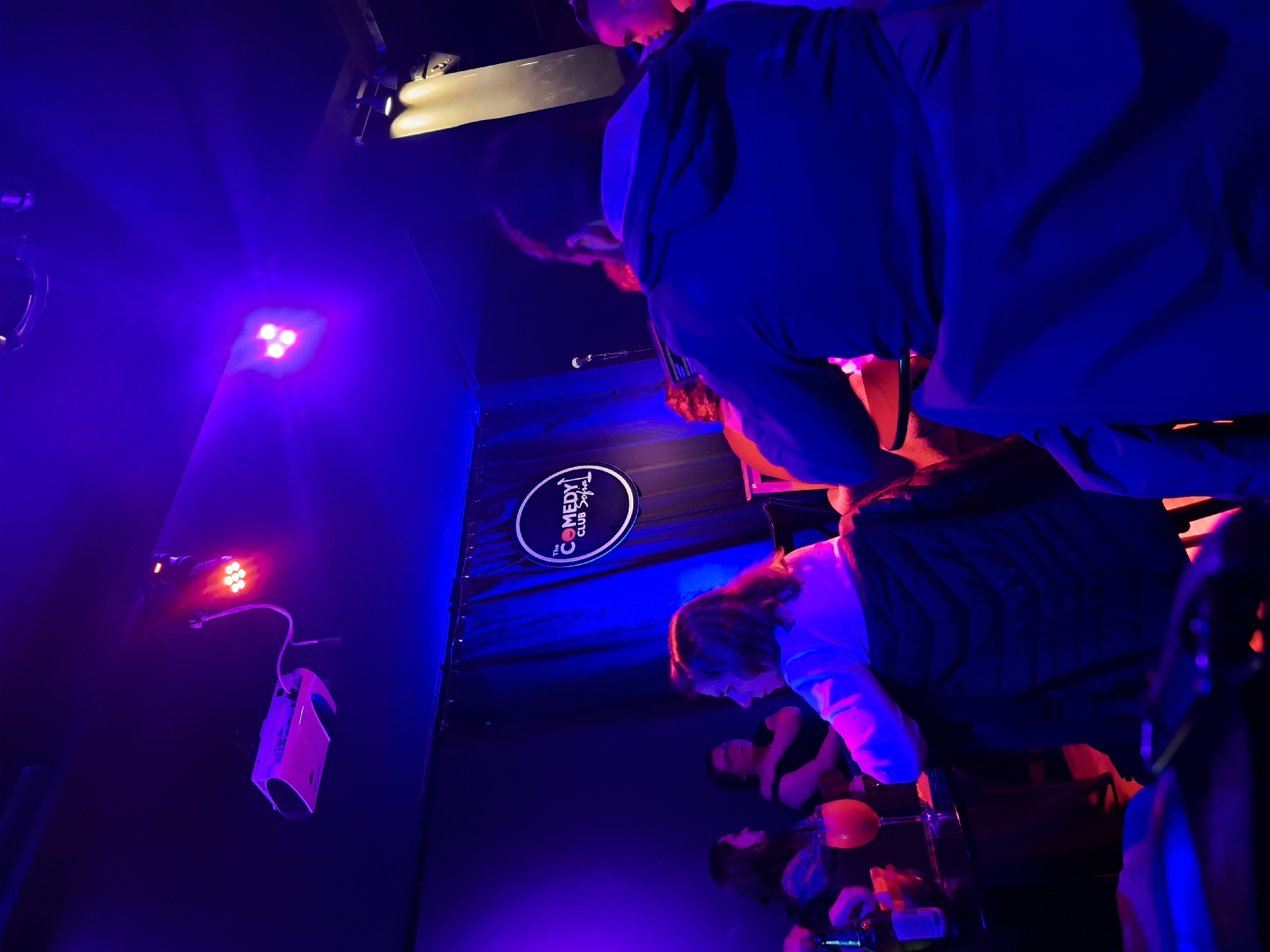 The image captures an interior scene in Sofia, Bulgaria, likely within a comedy club called "The Comedy Club Sofia". The image is rotated. The venue is dimly lit with blue and red stage lights and lighting equipment visible on the wall. There is a logo for the comedy club displayed on the back wall. Patrons are seated at tables, with drinks visible. People are in the foreground and are mostly visible from the waist up.