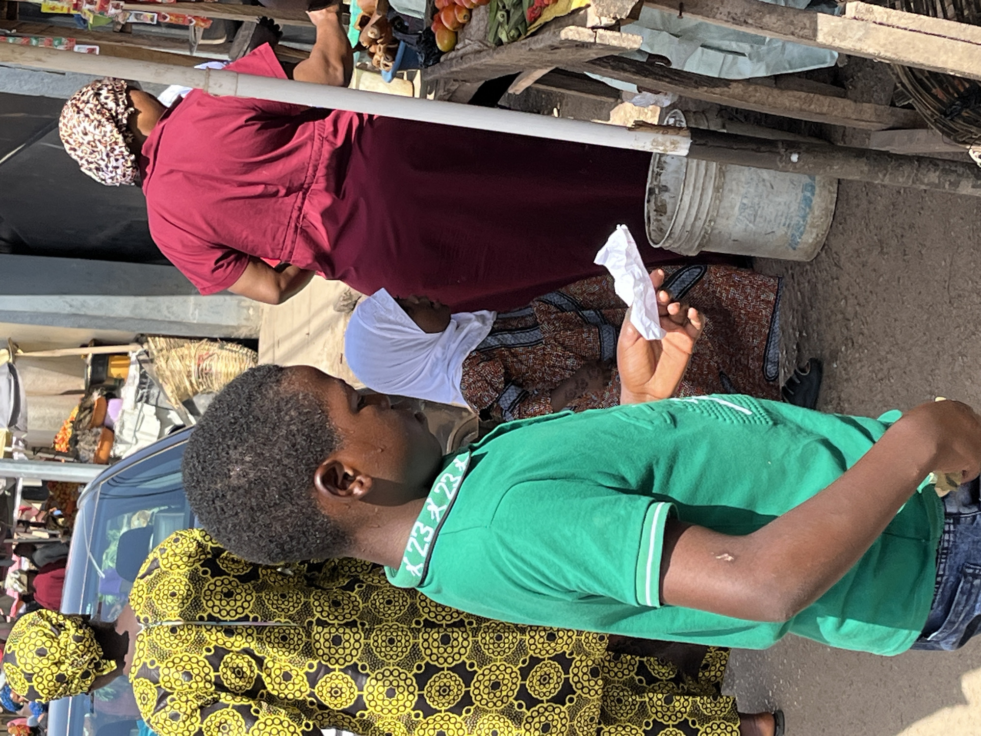 In an outdoor setting in Jos, Nigeria, a group of people are situated near a market stall. A young boy in a green polo shirt stands in the foreground, holding a piece of white paper. Beside him, a woman in a yellow and black patterned garment is partially visible. Further back, a woman with a white hijab sits near another woman in a red outfit and head covering. Behind them, a wooden market stall displays fruits and vegetables. A white bucket sits on the ground nearby.