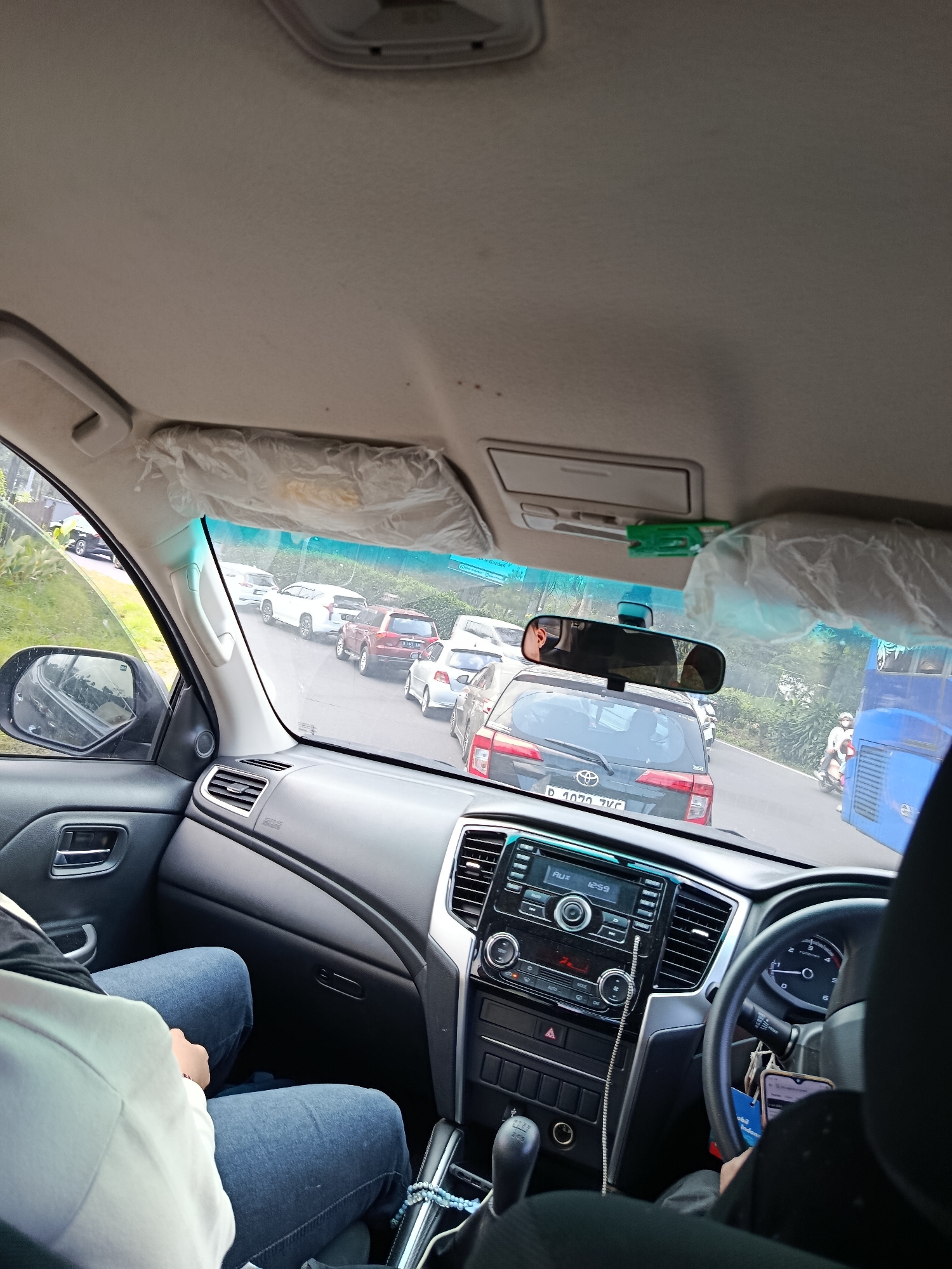 This image is taken from the interior of a car in Cisarua, Indonesia, looking forward through the windshield and out the passenger side window. The vehicle appears to be stuck in traffic on a two-lane road lined with lush green vegetation and trees. Several cars are visible in front of the subject vehicle, including a white SUV, a red SUV, and a black hatchback with the license plate "D 1070 JVC". A blue bus is in the right lane, and a person on a motorcycle is passing the bus. The car's dashboard, steering wheel, gear shifter, and radio are visible. The roof lining of the car shows signs of wear and tear, with some plastic sheeting visible, possibly for sun protection. The image suggests a hot and humid day, common in the region. The overall scene depicts a typical traffic situation in a scenic, possibly mountainous or hilly, area of Cisarua.