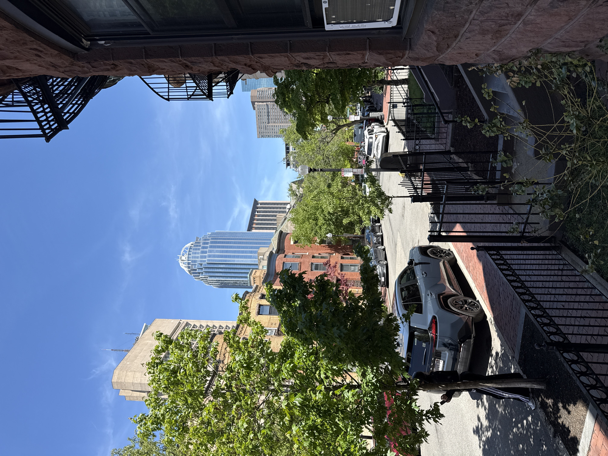The image depicts a street scene in Boston, Massachusetts, USA, viewed from an elevated position, possibly a window or balcony. The perspective is skewed. Tall buildings dominate the skyline, including the Prudential Tower with its distinctive dome. Various other buildings of different heights and architectural styles are visible along the street. The street is lined with parked cars and trees with green foliage. A section of a black wrought iron fence runs along the sidewalk on one side of the street. Sections of brick buildings and a fire escape are visible at the top of the frame. A person dressed in black, wearing sneakers with white stripes, is seen in the bottom right area of the image, partially obscured by a tree.