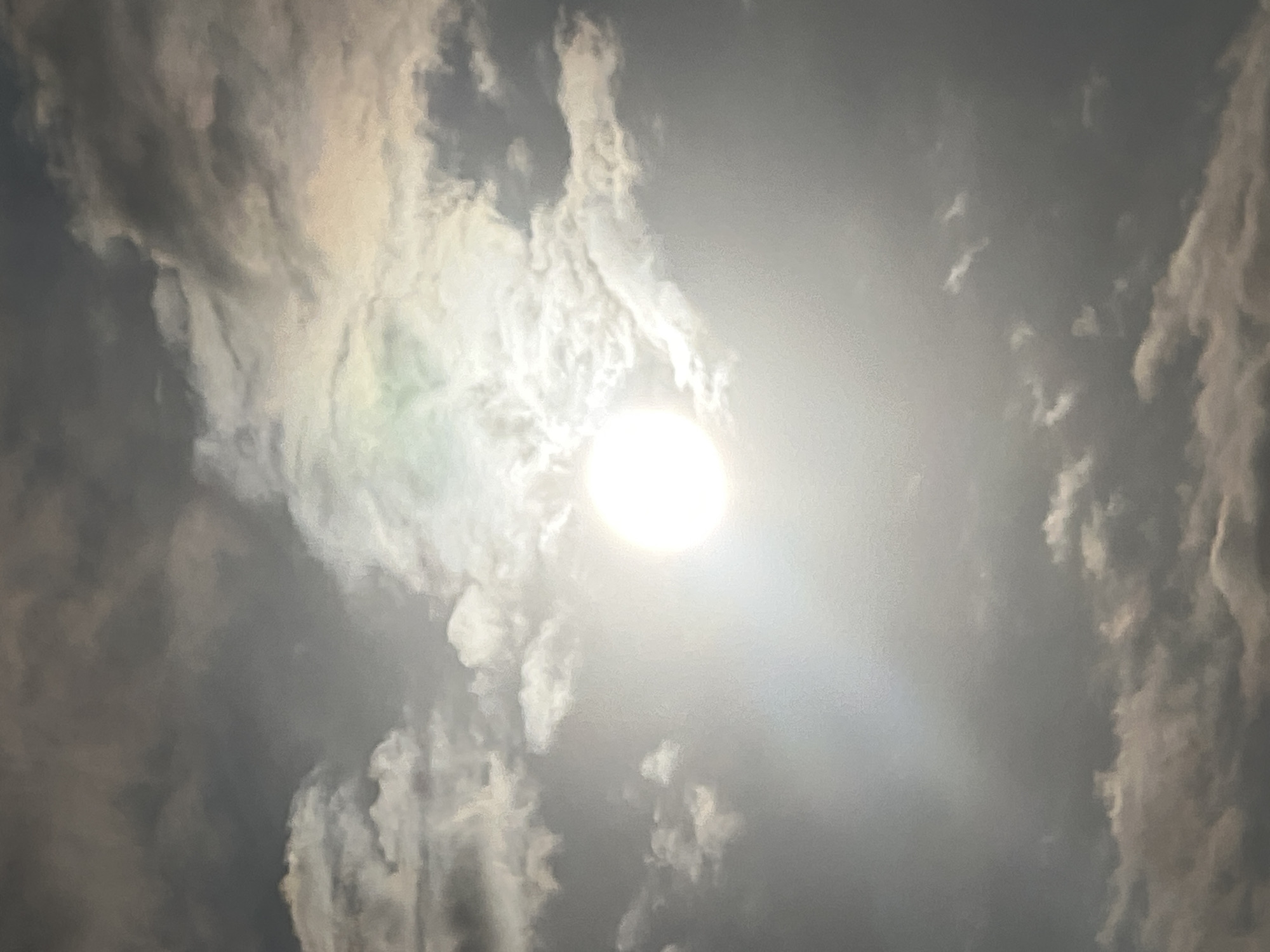 The image shows a bright sun partially obscured by scattered clouds in the sky. The sun is a luminous white circle, with a soft glow radiating outwards, suggesting it might be late afternoon or early morning, or that the clouds are thick. The clouds themselves are a mix of light gray and white, with some darker, more ominous patches visible, indicating potential for rain or stormy weather. The overall lighting is diffused, giving the scene a somewhat hazy or ethereal quality. There are no discernible objects, people, or specific landmarks in the image, making it impossible to determine a precise location within Kuta, Indonesia, or any other city. It is a purely atmospheric image of the sky.