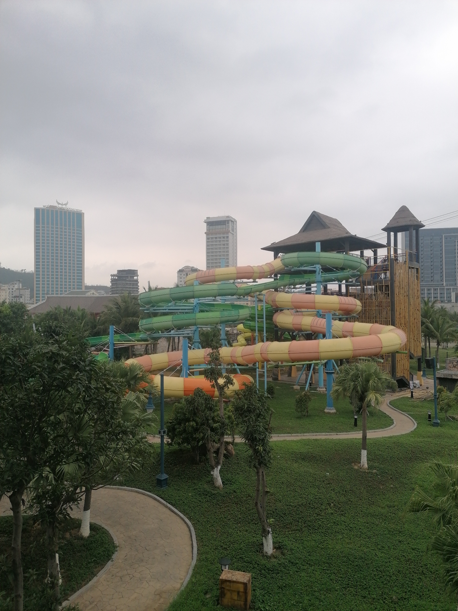 The image depicts a water park in Bai Chay Ward, Vietnam, under a cloudy sky. A large, multi-colored water slide structure, primarily green, yellow, and orange, dominates the scene. It is supported by blue columns and towers above a grassy area with scattered trees, including palm trees. A paved pathway curves through the park. In the background, several high-rise buildings are visible, typical of an urban setting. A wooden tower structure with a thatched roof stands next to the water slides.