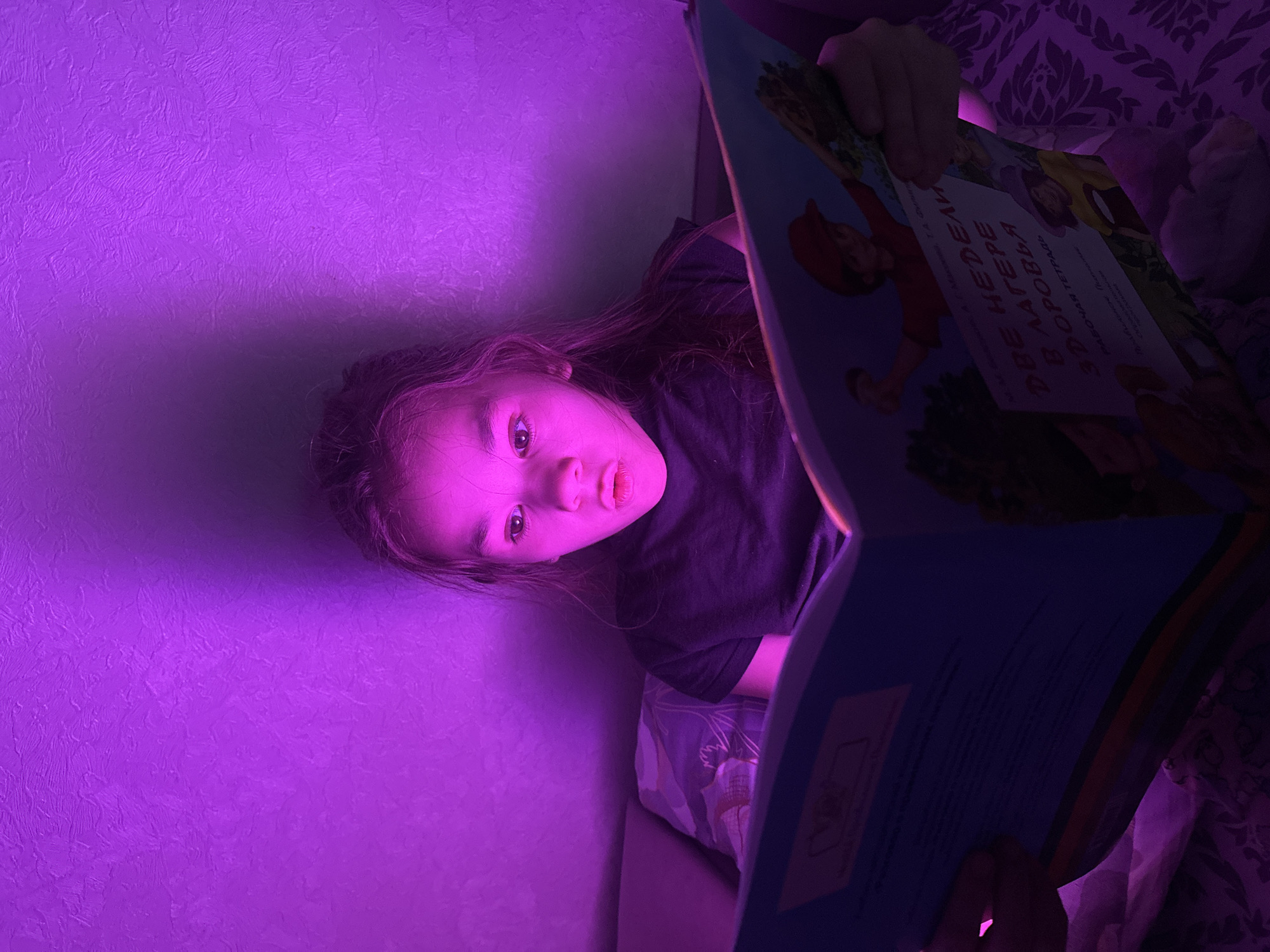 A young girl is depicted indoors, lying down and holding an open book, illuminated by a strong purple light. Her head is positioned towards the left side of the frame, and she looks directly at the viewer with a slightly parted mouth and a curious or neutral expression. She has long, dark hair and is wearing a dark t-shirt.
The background primarily consists of a textured wall, glowing vividly in purple light. To the right, glimpses of patterned bedding or a pillow are visible. The purple illumination strongly suggests it is nighttime or evening, and the primary light source is artificial, possibly an LED light strip.
The girl holds the book open, with both hands visible, though one is mostly obscured by the page. The visible page features illustrations of children and nature, and contains text in Cyrillic. The main visible title on the book cover reads: "ДВЕ НЕДЕЛИ В ЛАГЕРЕ ЗДОРОВЬЯ" which translates to "TWO WEEKS IN A HEALTH CAMP". Below that, "РАБОЧАЯ ТЕТРАДЬ" is visible, meaning "WORKBOOK". Further text includes what appear to be author names: "А.П. МАШУКОВА", "И.М. ДЕДЕНКО", and "Л.Ю. КАЙМАКОВ". The book cover itself is blue with colorful illustrations.
The scene captures a quiet moment of reading or learning, with the dramatic lighting adding a unique atmosphere.