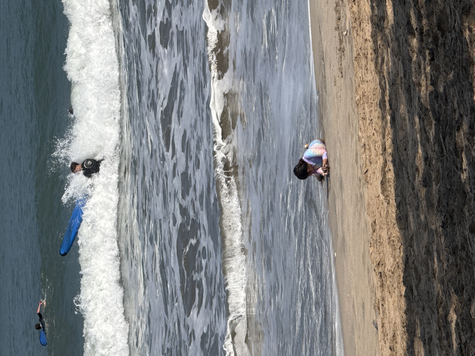 In Kuta, Indonesia, a child in a colorful swimsuit lies on the sandy beach, facing the ocean. In the background, two individuals are surfing. One surfer is near a blue surfboard in the water, while another surfer is closer to the shore, appearing to be interacting with a wave. The ocean exhibits moderate wave activity with whitecaps. The sand is textured with some footprints and shadows from overhead trees or structures.
