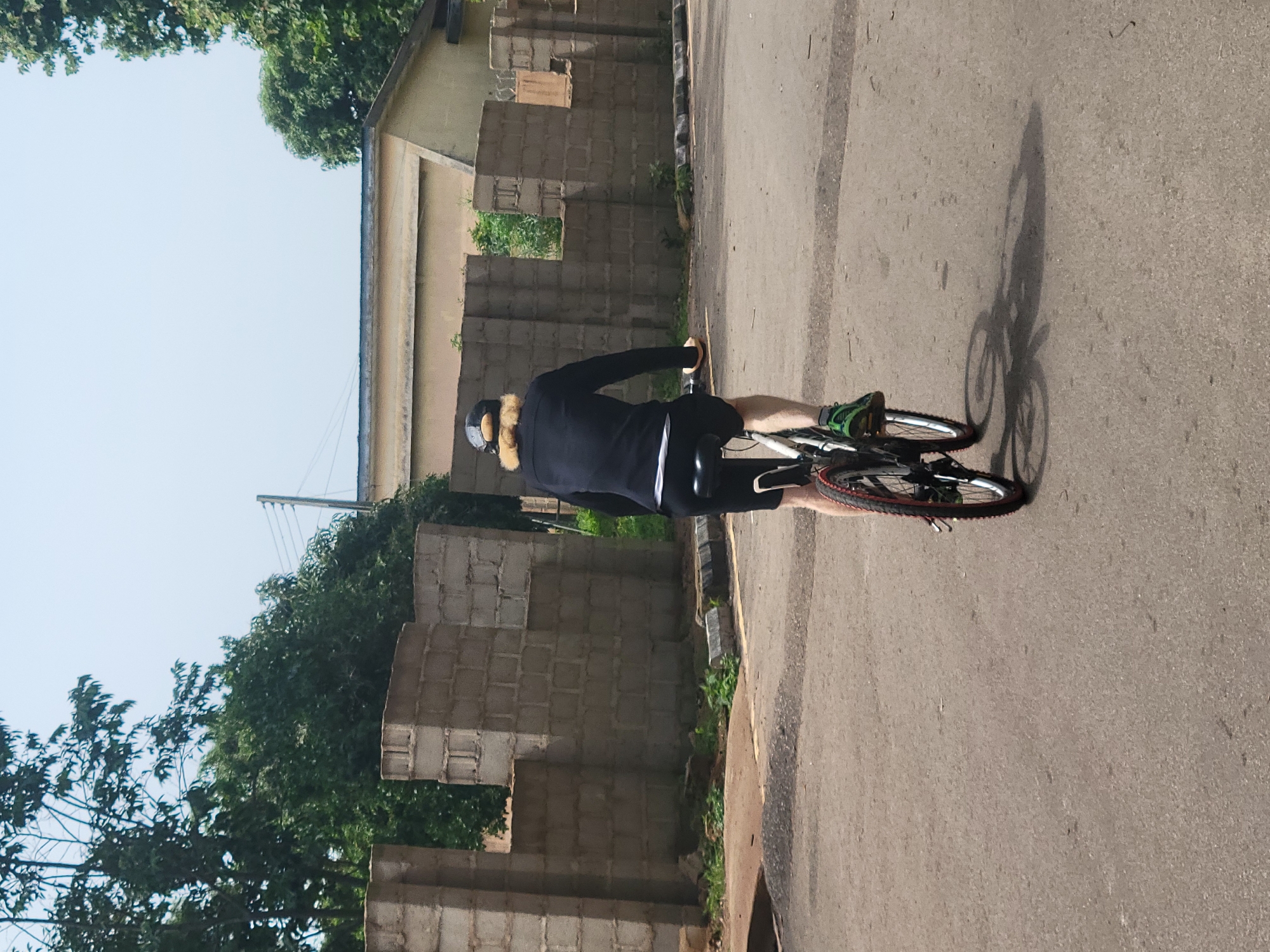 A person is riding a bicycle on an asphalt road, viewed from behind. The cyclist is wearing a black long-sleeved shirt, black shorts, a black baseball cap with a light-colored brim, and green athletic shoes. The bicycle has a white frame, black tires with red accents, and is casting a shadow on the road. The environment includes a grey concrete block wall on the left side of the road, overgrown green vegetation behind the wall, and a light-colored building in the background. Tall trees are visible above the wall. The scene appears to be in a semi-urban or developing area. The overall atmosphere is bright and sunny. The presence of the concrete block construction and the style of the building suggest this location could be within the city of Jos, Nigeria, where such construction and architecture are common.