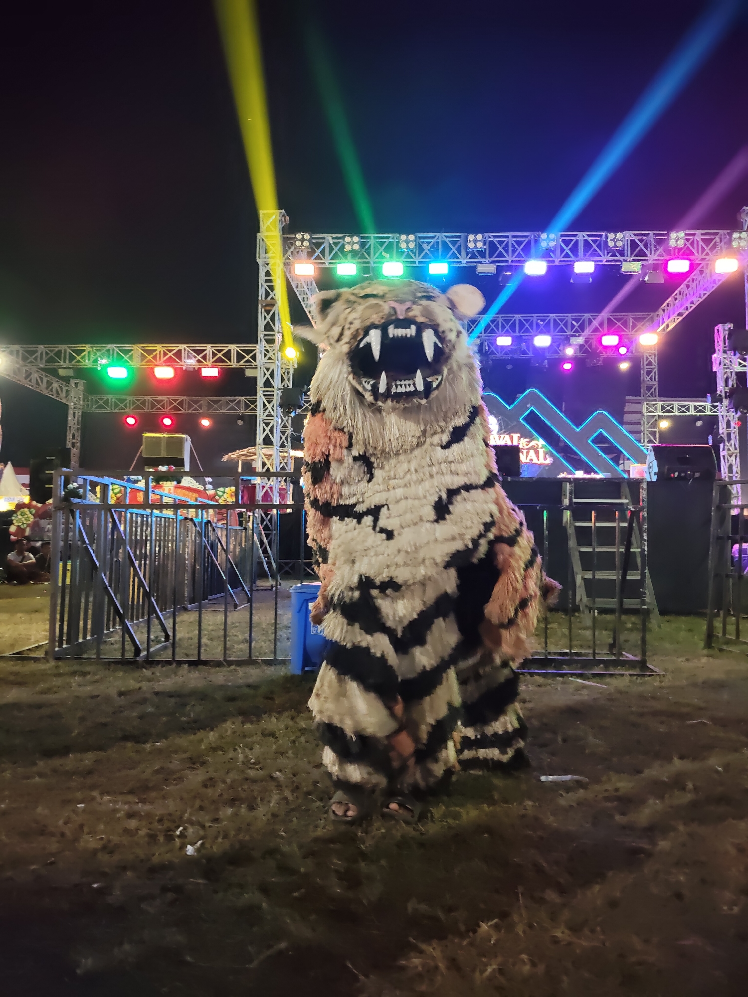 An individual is dressed in a tiger costume standing in front of a stage setup at night. The costume is a full-body suit with black and white stripes, a furry mane, and a large, fierce-looking head with visible teeth. The stage features a metal truss system with various colored lights, including green, red, yellow, blue, and pink, casting beams into the night sky. In the background, there's a large neon sign with stylized letters, possibly indicating a festival or event. A metal barricade separates the foreground from the stage area. The ground is covered in dry grass and some scattered debris. A blue trash can is visible near the barricade. The location is identified as the city of Sumenep, Indonesia, suggesting this is likely a public event or performance.