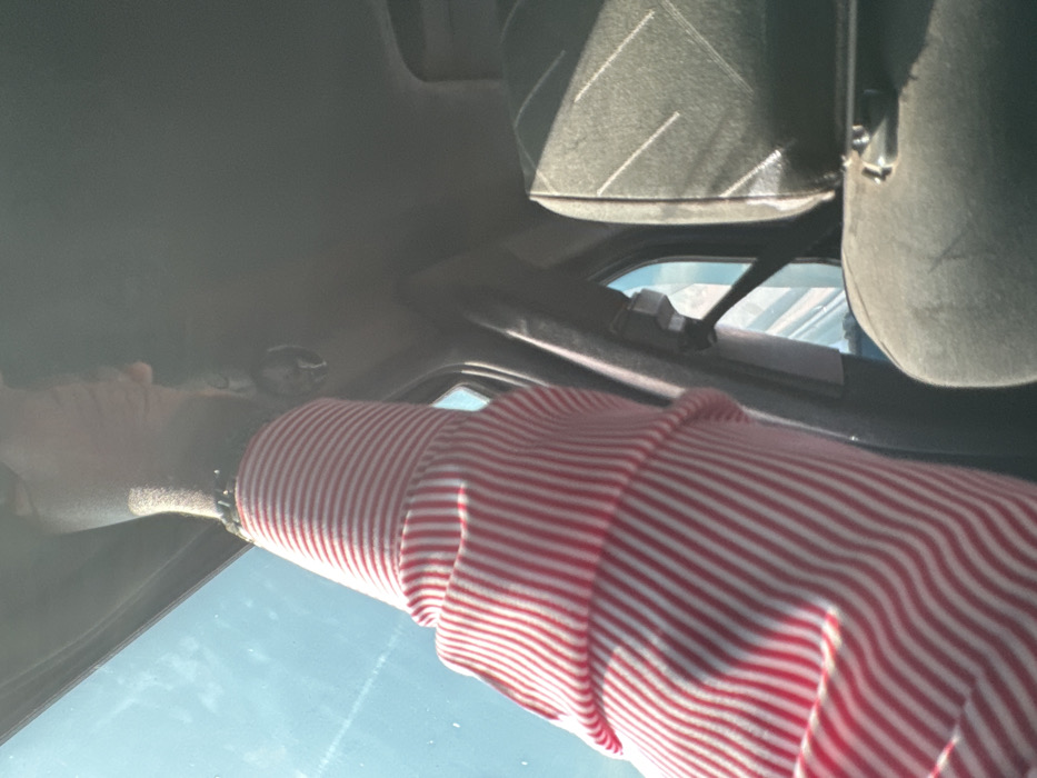 The image depicts the interior of a vehicle, presumed to be in Dokwa, Nigeria. A dark-skinned arm, dressed in a red and white vertically striped long-sleeved shirt, extends upwards towards the vehicle's light gray headliner. A dark beaded wristband is visible on the wrist, from which a small, dark, decorative object (possibly a skull figurine) hangs. Through the vehicle's window, a clear blue daytime sky is visible to the left. On the right, a dark gray car seat with faint diagonal striped patterns is partially visible, illuminated by direct sunlight. A black seatbelt and its attachment mechanism are also present on the dark gray pillar to the right of the arm.