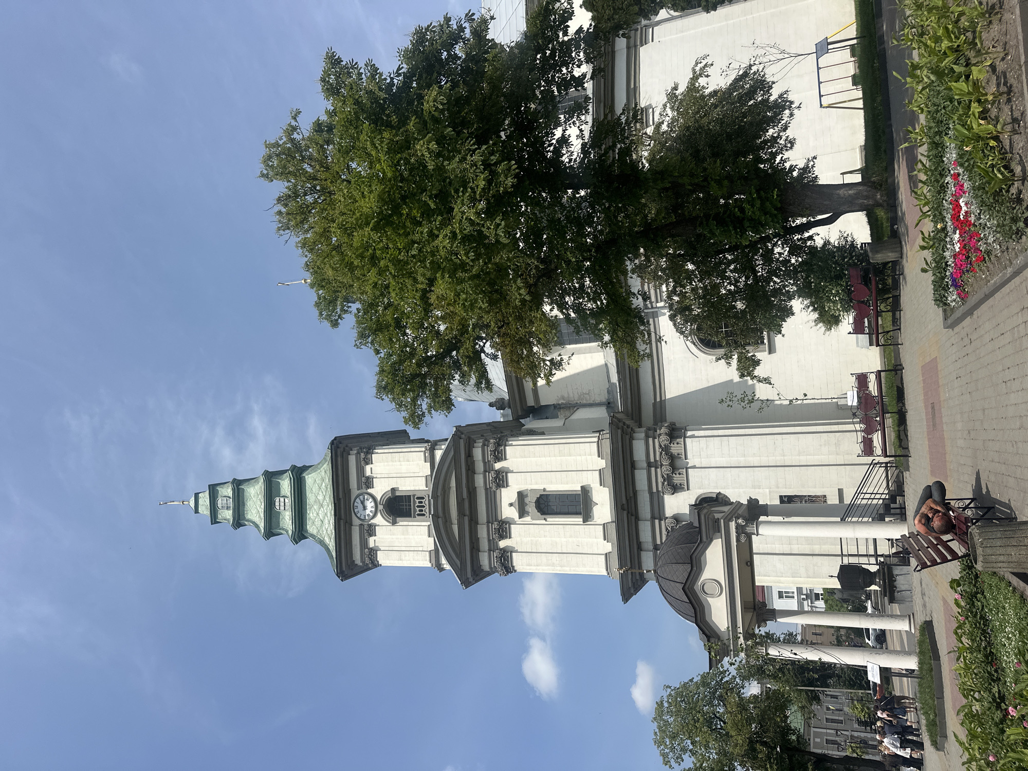 The image shows the Ternopil Church of the Resurrection of Christ, a prominent landmark in Ternopil, Ukraine. The church features a tall, white tower with a green spire and a clock face. A large, leafy tree stands in front of the church, partially obscuring its facade. In the foreground, there is a public square with benches and flower beds. One bench is occupied by a person sleeping. Several other people are visible in the background, walking near the entrance of the church. The sky is blue with scattered clouds.