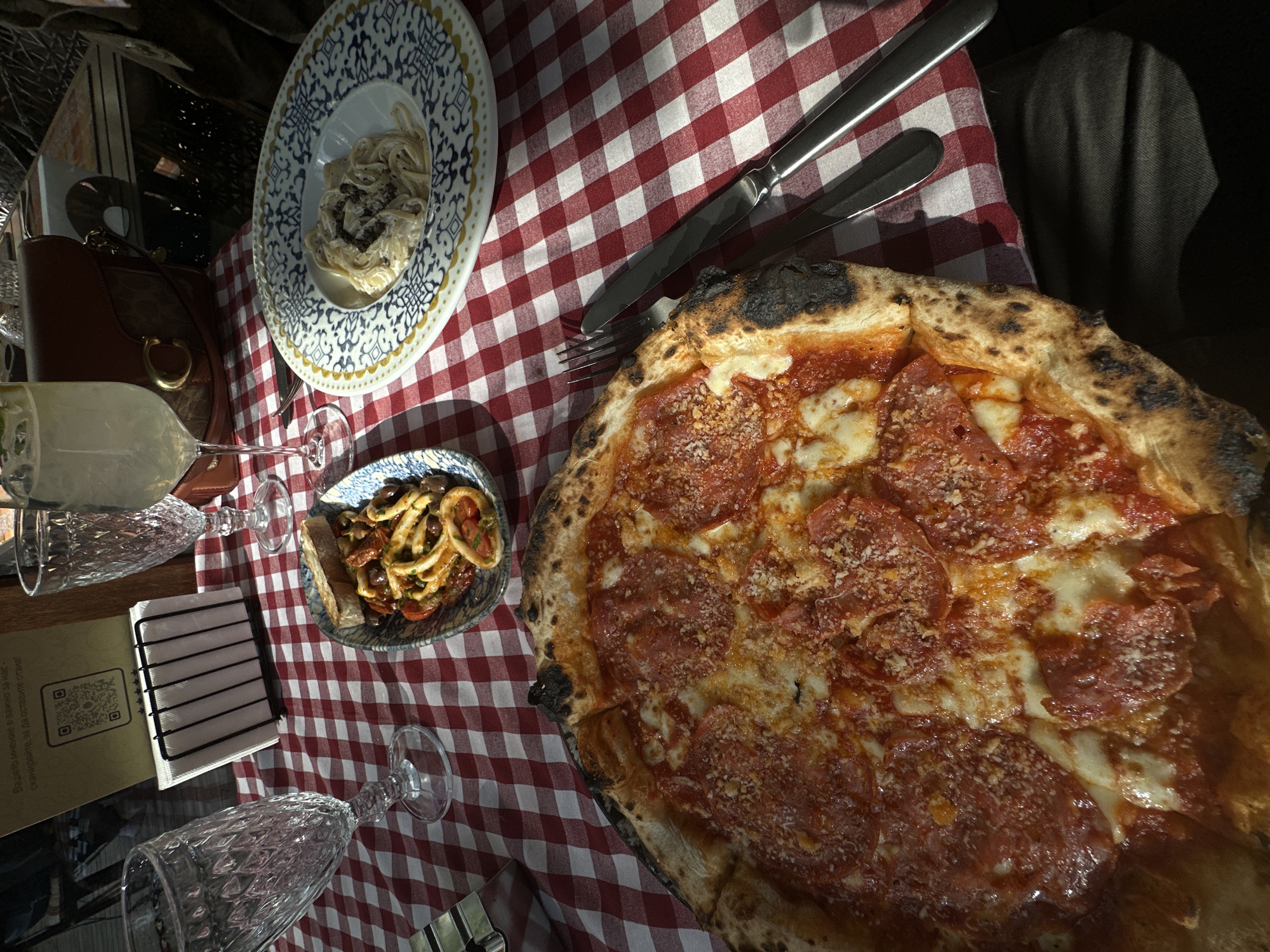 A table covered with a red and white checkered tablecloth is laden with food and drink. A pepperoni pizza with a charred crust dominates the right side of the frame. To the left of the pizza are a fork and knife. Further left is a blue patterned bowl containing calamari or a similar seafood dish. A patterned plate with what appears to be pasta is above the bowl. To the left of the bowl is a wine glass and a napkin holder. A handbag is visible in the upper left corner. A second wine glass is at the bottom of the frame. A folded menu card is present near the napkin holder, featuring a QR code and text. The scene suggests a restaurant setting, but the precise location in Sofia, Bulgaria, cannot be determined without additional information.