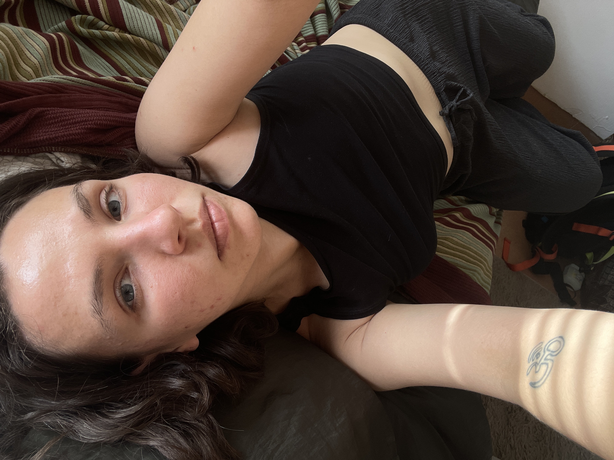 A woman with fair skin is lying on a bed with a striped comforter. She is wearing a black tank top and dark pleated pants. She has dark hair and grey eyes. The woman's right arm is extended, revealing a tattoo on her inner forearm. The scene appears to be indoors, possibly a bedroom. Budva, Montenegro is not indicated in the image.