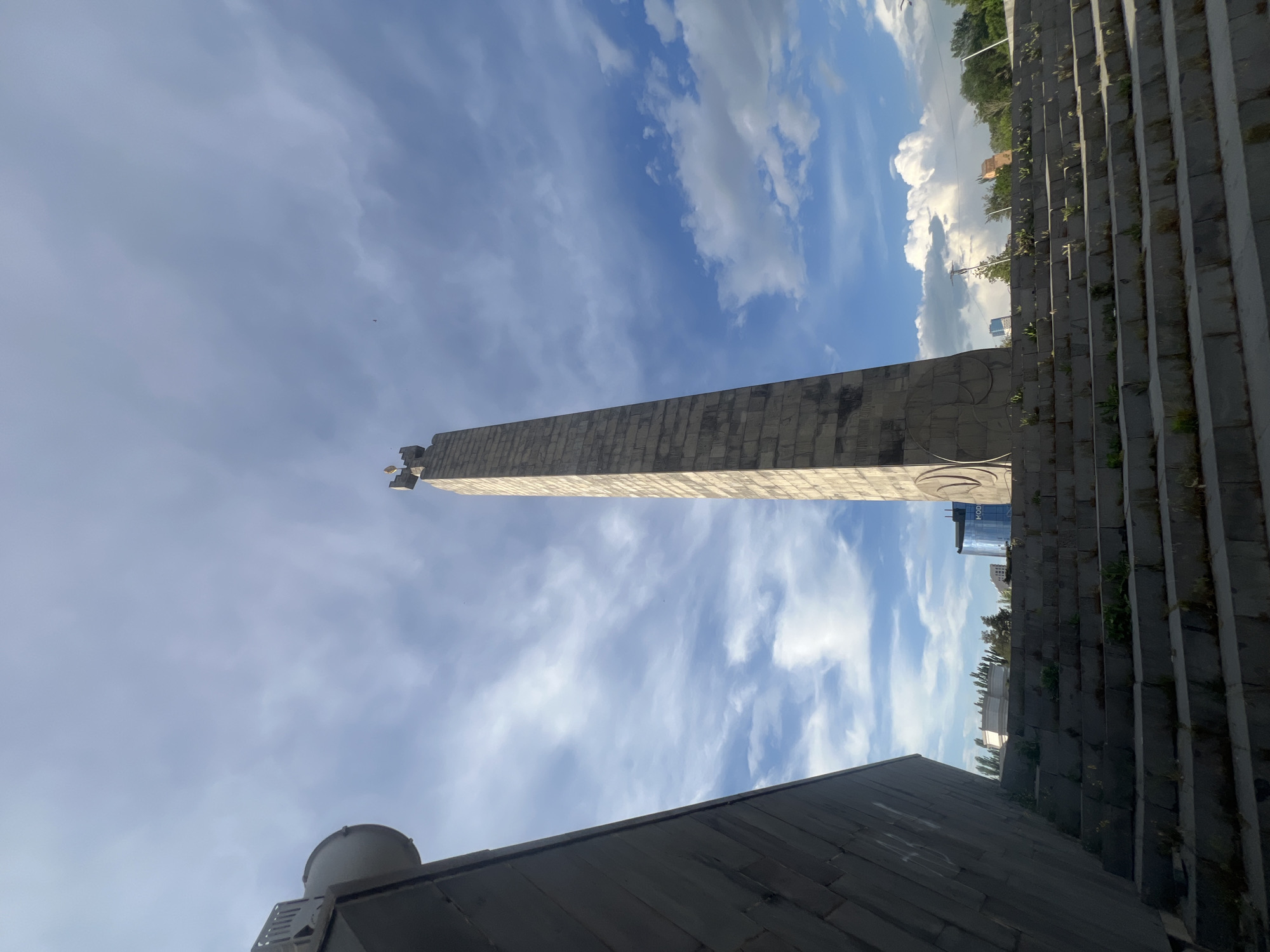 The image captures a wide-angle, low-angle view of the "Reborn Armenia" stele at the Armenian Genocide Memorial Complex (Tsitsernakaberd) in Yerevan, Armenia.
A tall, grey, stone-clad obelisk, slightly off-center to the right, extends vertically from the mid-ground towards the upper edge of the frame. Its surface exhibits a textured or block-like pattern, and a stylized golden element, possibly a multi-pronged sun or flame, crowns its apex. To the left, a large, dark grey, angular concrete structure forms a steep diagonal plane, partially obscuring the left sky and a portion of the steps. A large, light-colored, circular object, potentially a speaker or light fixture, is partially visible at the top-left edge of this structure.
In the foreground, a wide set of grey stone steps, marked with small patches of green vegetation growing between the risers and treads, ascends towards the background. Beyond the steps, various urban elements are visible: trees line a distant ridge, and a modern building with a reflective blue facade is discernible to the left of the stele. The sky above is bright blue with scattered white and grey cumulus and cirrus clouds. No individuals are present in the scene.