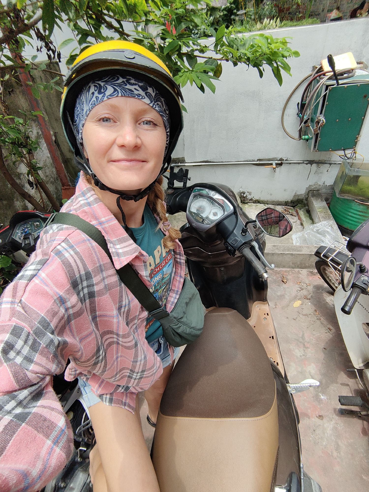 A woman wearing a yellow helmet, a blue patterned bandana, and a pink plaid shirt takes a selfie while standing next to a black scooter in Bai Chay Ward, Vietnam. She has a green fanny pack across her chest. The scooter has a brown seat. Behind the scooter, another motorcycle is visible. In the background, a white wall with green electrical equipment and pipes is seen, along with green foliage. The ground is concrete.