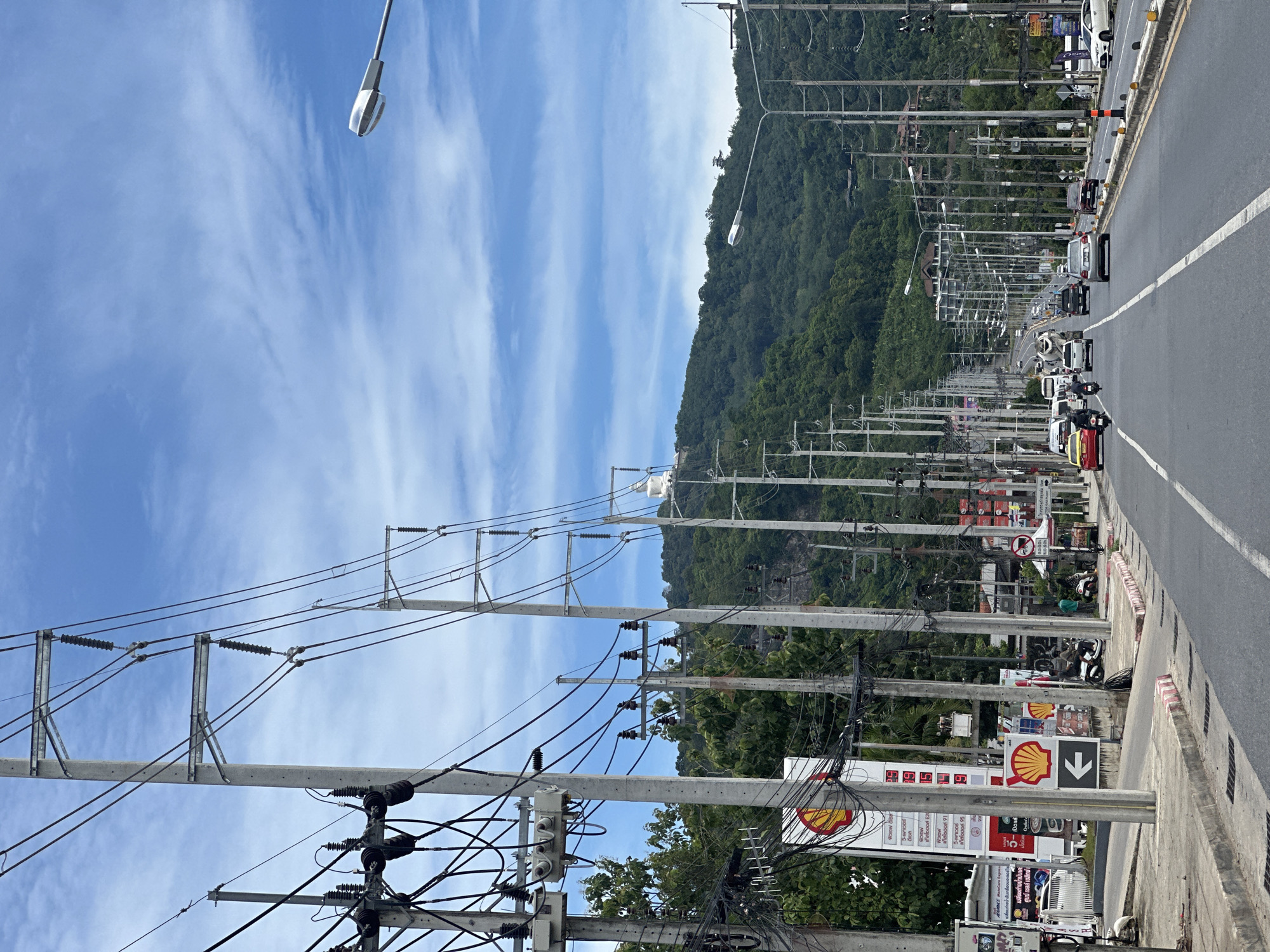 The image depicts a wide, multi-lane road stretching into the distance, lined with numerous utility poles carrying a dense network of wires. On the left side of the road, there is a sidewalk with a Shell gas station visible, featuring prominent signage and a white arrow pointing left. A few motorcycles and people are present on the sidewalk. Vehicles, including cars, a taxi, and a bus, are driving on the road. The road is bordered by a grassy verge and a dense, green, tree-covered hillside in the background. The sky is blue with scattered white clouds. A large white statue of Buddha is visible on the hillside in the distance, atop the trees. Streetlights are mounted on poles along the road. The scene appears to be a busy thoroughfare in a tropical or subtropical region.