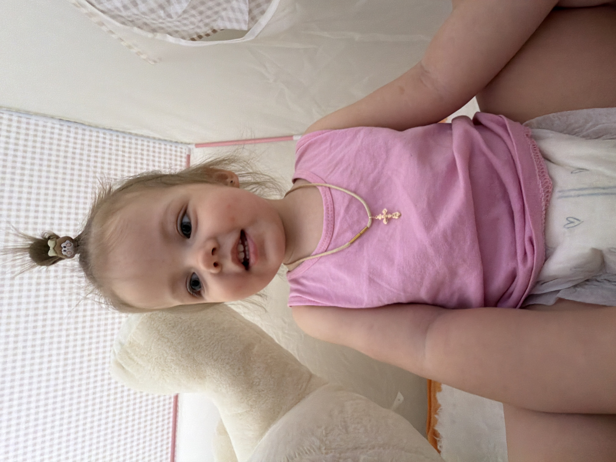 An overhead portrait captures a young child, likely a toddler, looking directly at the camera with a playful expression, a slight smile revealing a few teeth. The child has light-colored hair pulled into a small, spiky ponytail adorned with a small, character-themed hair tie. Around their neck, a light cord holds a small golden cross pendant. They are wearing a plain pink sleeveless top and a disposable diaper with faint blue heart patterns visible.
The child appears to be sitting or leaning against a soft, light-colored plush toy or pillow, visible to their left. The setting is indoors, featuring a background of light-colored material with a subtle checkered or gingham pattern, suggesting a play tent or an enclosed, fabric-lined space. Above the patterned area, a plain light surface, possibly a ceiling or another fabric panel, is visible. A thin pink rod or structural element can be seen in the background, reinforcing the idea of a soft enclosure.
The lighting is soft and even, indicating an indoor environment, likely during the day. The child's direct gaze and happy demeanor suggest a candid or interactive moment. There is no visible text in the image. The scene conveys a sense of innocent playfulness in a comfortable, child-friendly environment, possibly in Reshetnikovo, Russia.
