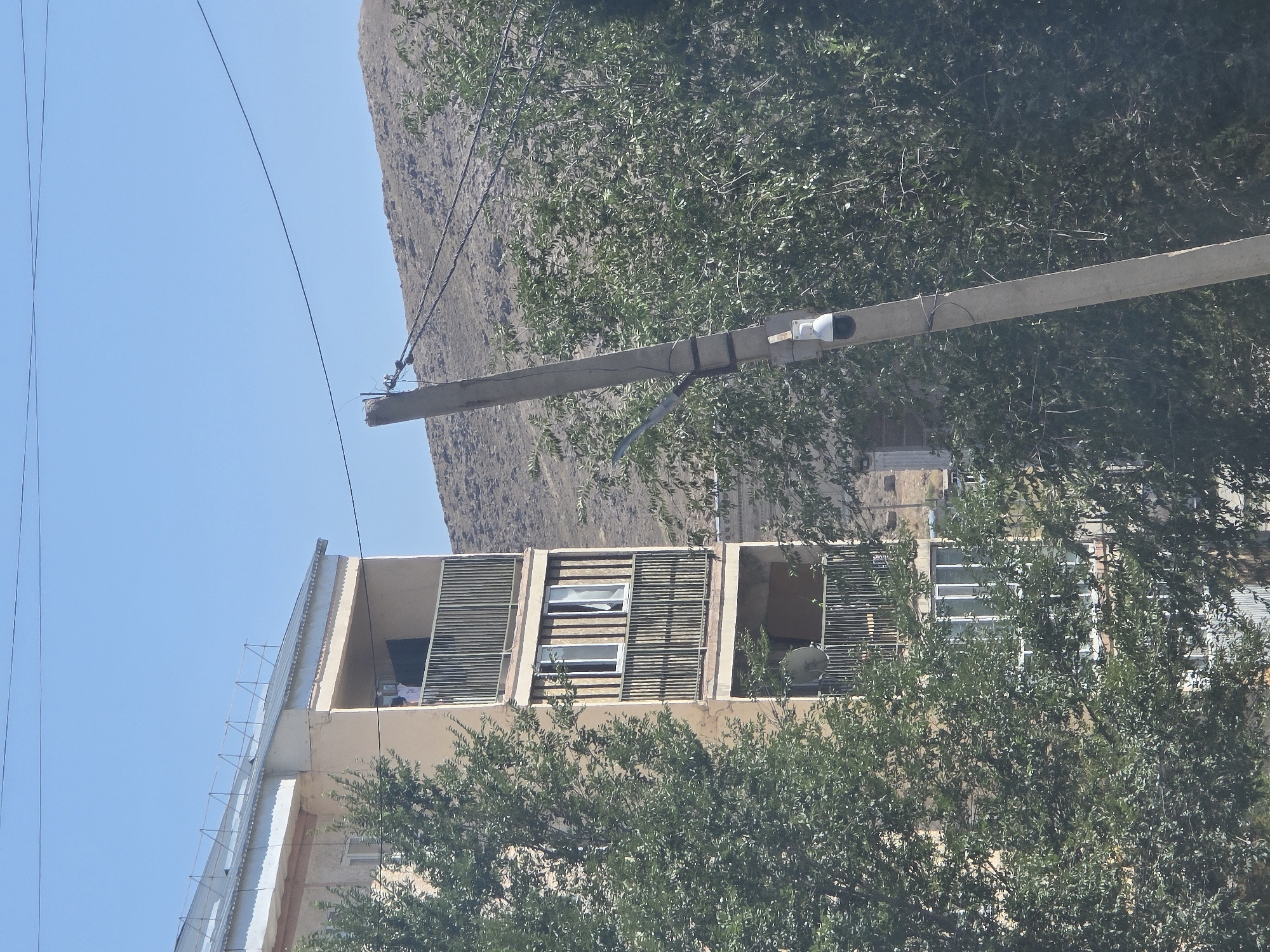 The image shows a concrete utility pole with a security camera attached, positioned in front of a multi-story apartment building and a tree-lined background. The apartment building is light tan with multiple balconies and windows. A person is visible on one of the balconies. The sky is clear blue, and a rocky, barren hillside is visible in the distance. The scene appears to be in a residential area, with the utility pole and camera suggesting surveillance or infrastructure presence. The vegetation is dense and green, indicating a temperate climate or season. The specific location within Qaratau, Kazakhstan, is not explicitly identifiable from the visual information presented.