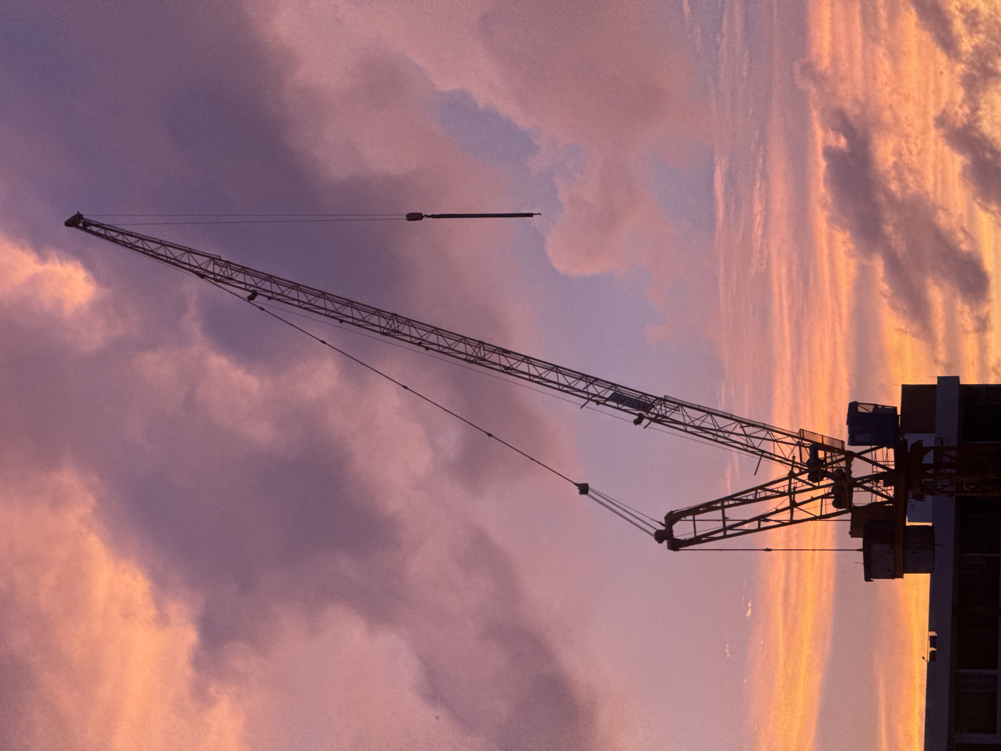 A construction crane is silhouetted against a dramatic sunset sky filled with pink and orange clouds. The crane, a tall, lattice-like structure, extends diagonally from the lower left to the upper right of the frame. At the base of the crane, the top of a building or structure is visible, also in silhouette. The sky is a prominent feature, with swirling clouds in various shades of pink, purple, and orange, indicating either sunrise or sunset. There are no people or animals visible in the image. The scene suggests an active construction site in Karon, Thailand, during twilight.