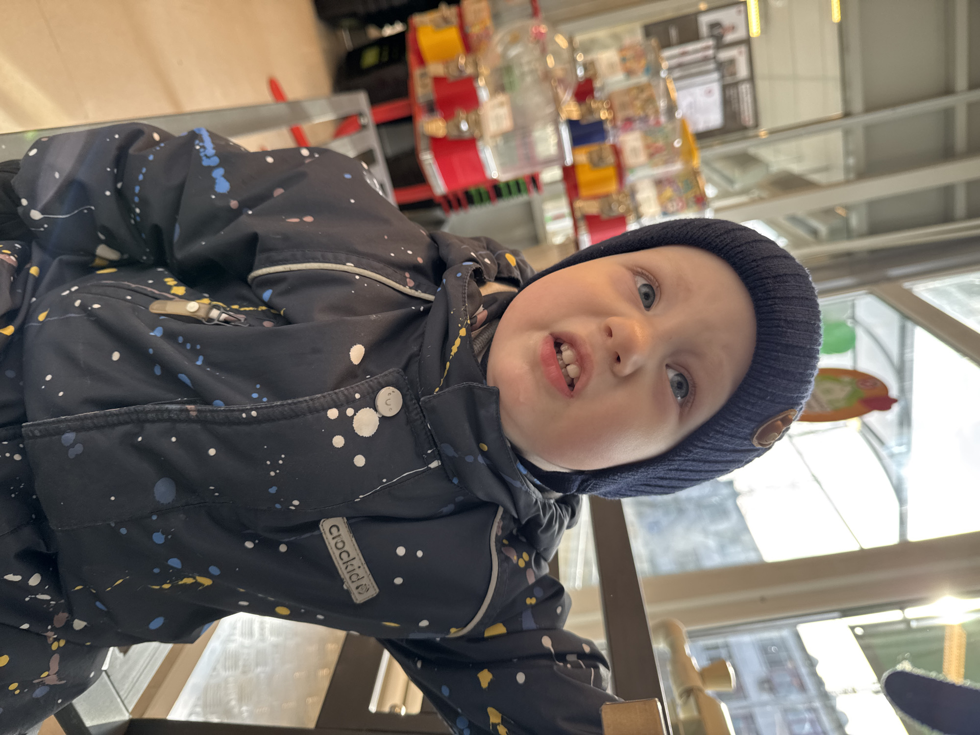 A low-angle, rotated close-up shot features a young child, likely a toddler, looking directly into the camera with a playful or curious expression, showing some teeth. The child has light-colored eyes and is wearing a dark blue knitted beanie and a dark blue winter jacket. The jacket is adorned with a pattern of white, blue, and yellow paint splatters. A white rectangular label with the brand name "crockid" is visible on the child's right sleeve.
The background, out of focus, suggests an indoor public setting, possibly a retail store or supermarket, in Chaikovsky, Russia. Blurred shelving units with various small, colorful items, potentially products like candy or postcards, are visible. Bright natural light streams in from large windows, indicating it is daytime. A metallic or wooden structure, possibly a railing or part of a display, is present in the foreground near the bottom of the image. The overall mood conveyed by the child's expression is one of engagement and cheerfulness.