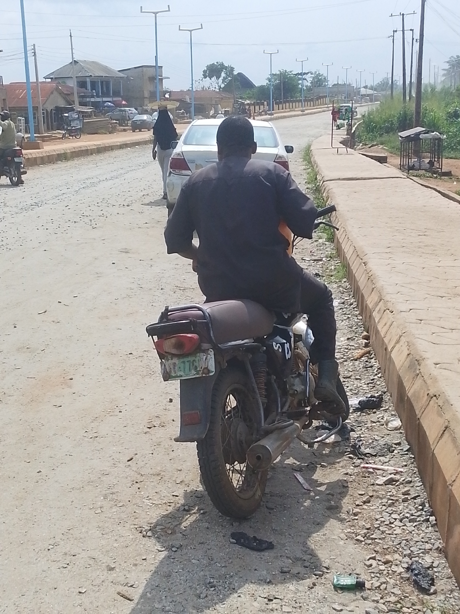 The image shows a man from the rear riding a motorcycle on a dusty, unpaved road in Abeere, Nigeria. The motorcycle is maroon and has a license plate. The man is wearing a dark shirt and black pants. To the left of the road are buildings, including a house with a corrugated metal roof and a white car. A person in a black hijab is walking on the side of the road. To the right of the road is a concrete sidewalk with a drainage ditch and some debris, including a green bottle and plastic bags. In the distance, there are streetlights and more buildings. The sky is clear and the sun is shining.