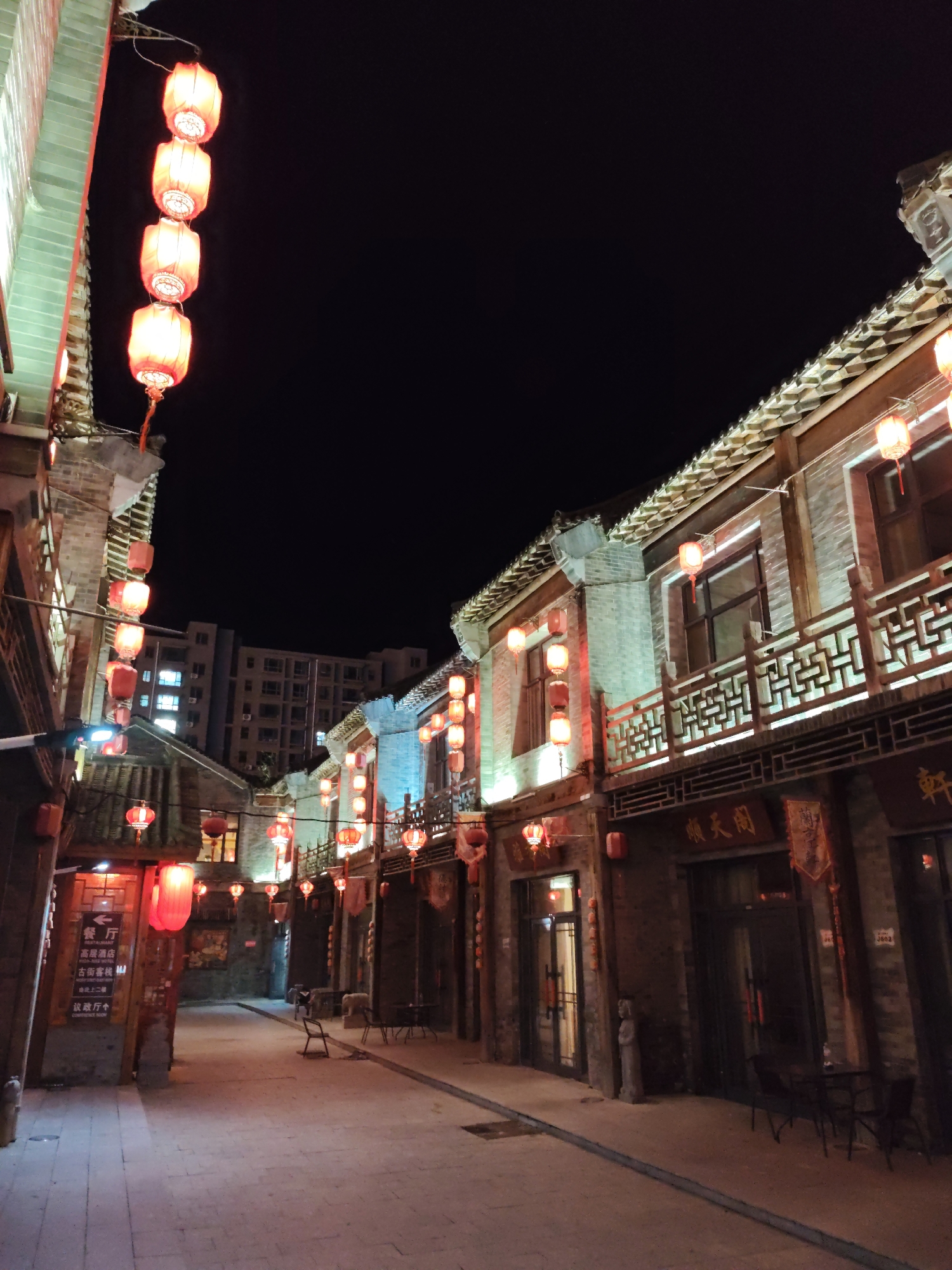 The image depicts a narrow street at night in Lindian, China, illuminated by numerous red lanterns. The street is lined with traditional brick buildings featuring ornate wooden balconies and tiled roofs. In the background, a modern, multi-story residential building stands out against the dark sky. The ground is paved with cobblestones. Several establishments are visible along the street, indicated by signs with Chinese characters. Red lanterns are strung vertically along the buildings and hang from poles, casting a warm glow on the scene. Chairs and tables are set up outside some of the establishments, suggesting outdoor dining or seating areas. There are no people visible in the image. The atmosphere is serene and evokes a sense of historical ambiance mixed with modern urban elements.